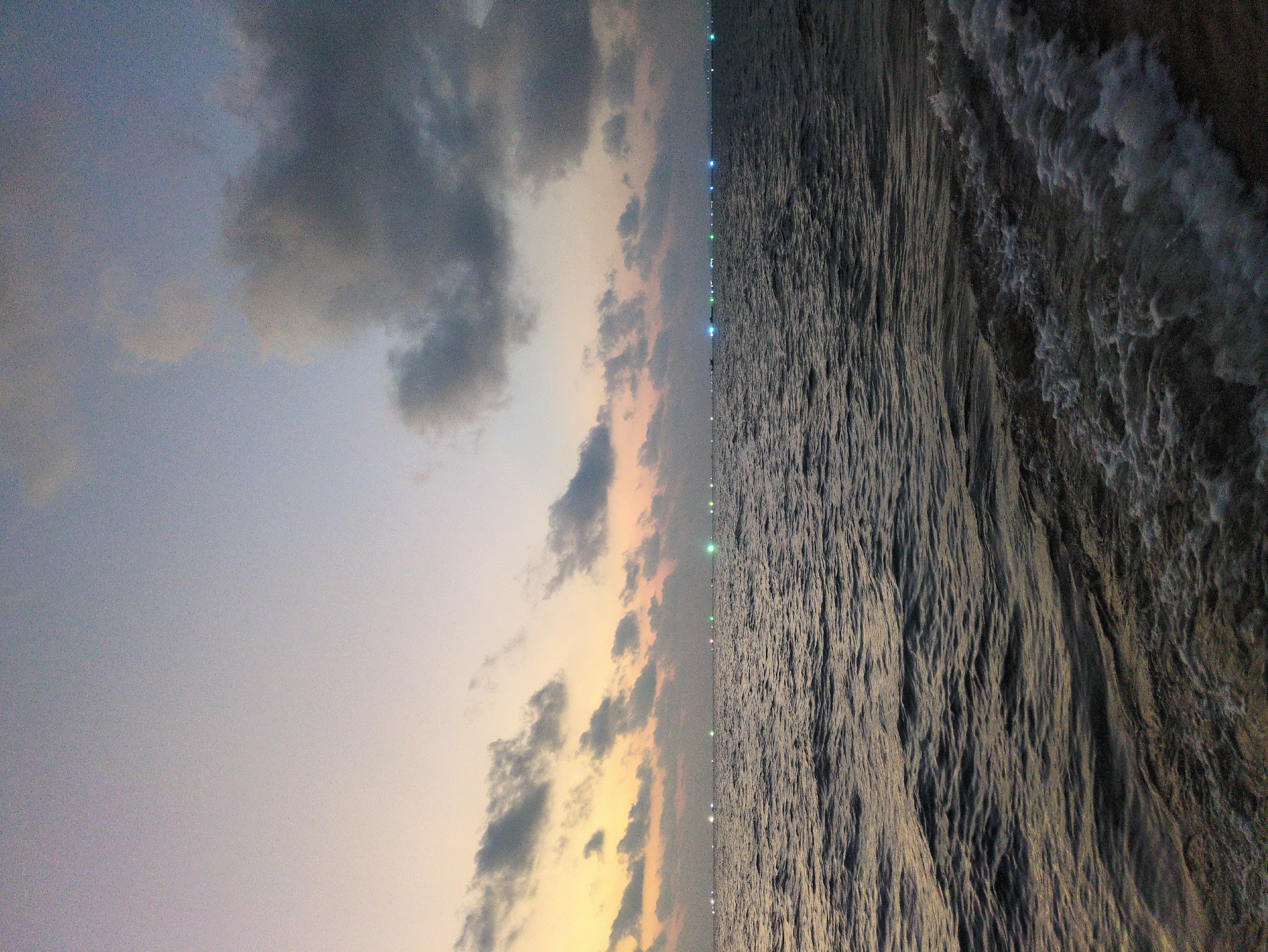 The image captures a twilight scene at a beach in Phú Quốc, Vietnam. The left side of the image displays the sky, featuring gray and beige clouds against a dusky backdrop. A gradient of color transitions from a light blue-gray to a yellow-orange near the horizon. Along the center, aligned vertically, are faint, twinkling lights appearing to be from distant structures or vessels at sea. The right side of the image is dominated by the dark, textured surface of the ocean, with visible wave patterns and foam. The perspective suggests the photograph was taken near the shore, looking out towards the sea. There are no visible people or other identifiable subjects.