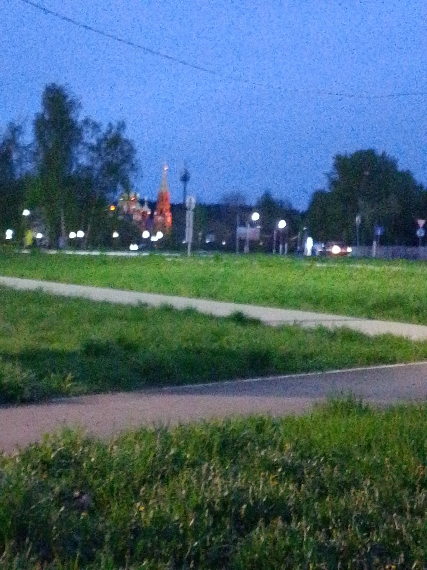The image shows an outdoor scene in Chaikovsky, Russia, at dusk. In the foreground, there is green grass and a gray asphalt path. Beyond the path is a field of grass with a lighter-colored walkway cutting through it. In the background, blurred trees and street lights are visible. The outline of a red and white tower is visible amongst the lights, a possible landmark of Chaikovsky. The sky is a dark blue, indicating the low light conditions. Some street signs can be seen alongside what appears to be a road with a vehicle with headlights on it.