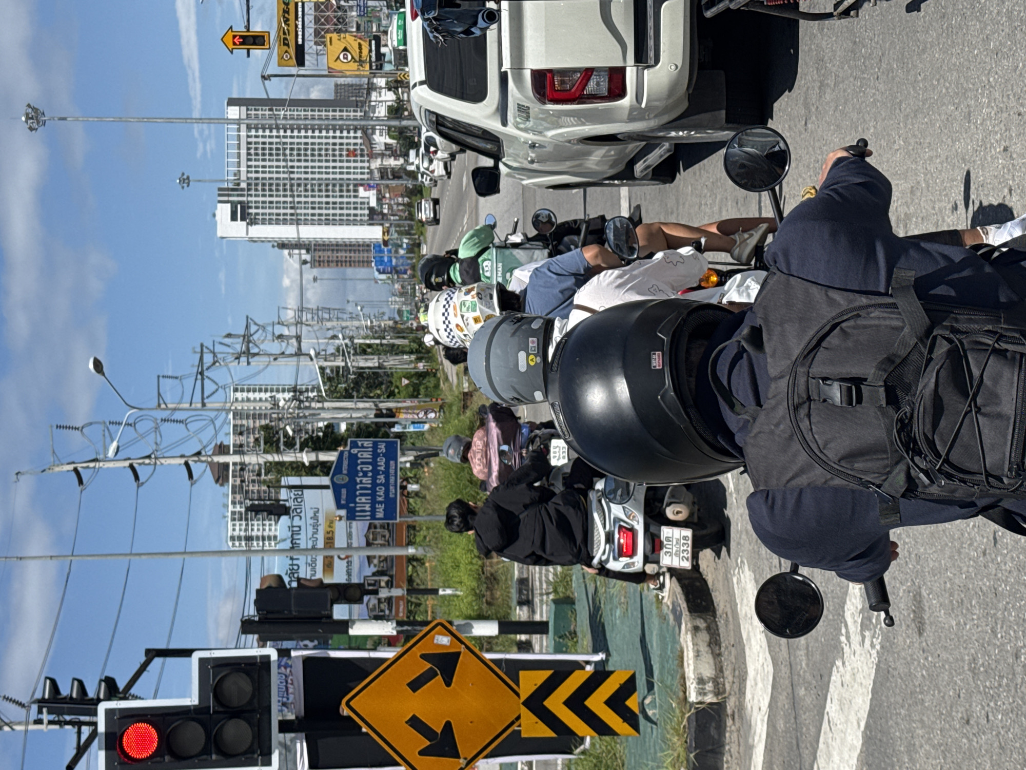 The image shows a street scene in a city, likely in Thailand. In the foreground, a person wearing a black helmet and backpack rides a motorcycle. Ahead of them, other motorcycles and a white pickup truck are visible in traffic. To the left, a traffic light shows a red signal, and a diamond-shaped yellow road sign with two black arrows pointing left and down is displayed. In the background, tall buildings, power lines, and other vehicles indicate an urban environment. The sky is blue with scattered clouds. The sign in the middle ground indicates "MAE KAO SA-AAD-SAI" and "INTERSECTION". The overall impression is a busy street with motorized traffic on a sunny day.