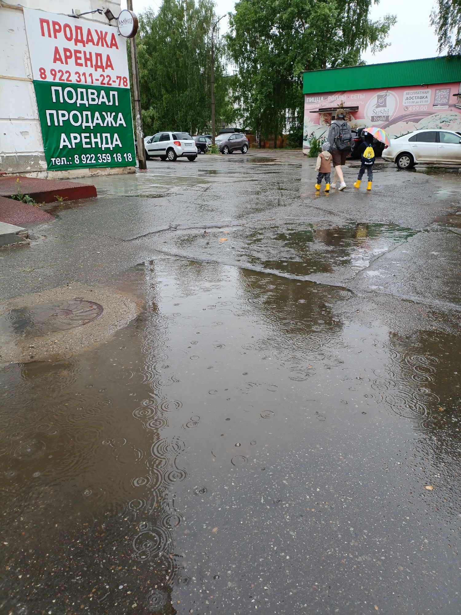 The image shows a rain-soaked street in Chaikovsky, Russia. Raindrops are visible on the waterlogged pavement, creating ripples. A manhole cover is seen near the foreground.
In the background, a woman is walking with two children, all wearing yellow boots. The children seem to be walking towards the right. A building with a sign that reads "Sushi Yoshi" in English can be seen. A restaurant menu visible on the building includes pizza, rolls, and other items. Cars are parked nearby.
A sign in Russian advertising property for sale or rent is on the left. The phone number "+7-922-311-22-78" is visible on the sign, along with "Podval Prodazha Arenda," which translates to "Basement Sale Rent."