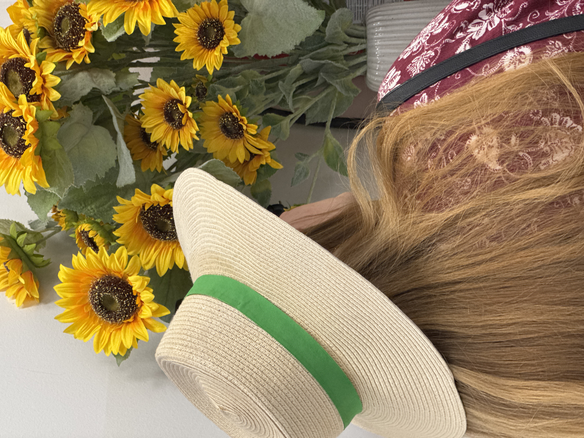 The image depicts a close-up, over-the-shoulder view of a person wearing a straw hat with a green ribbon, positioned in front of a large bouquet of artificial sunflowers. The person has long, blonde hair and is wearing a dark red dress with a white floral pattern. The sunflowers are vibrant yellow with dark centers, and they are arranged in a vase. The background appears to be a neutral-colored wall. The scene is indoors, and no specific location in Podgorica, Montenegro can be identified from the visual information provided.