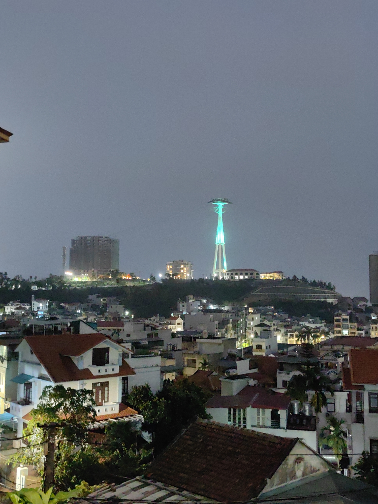 The image depicts a nighttime view of Bai Chay Ward, Vietnam, with a prominent tower illuminated in green at the center. The tower is situated on a hill among other buildings. In the background, a large, unfinished building stands out to the left of the tower. The foreground is dominated by houses with red tile roofs. The sky is overcast. There are no people visible in the image.