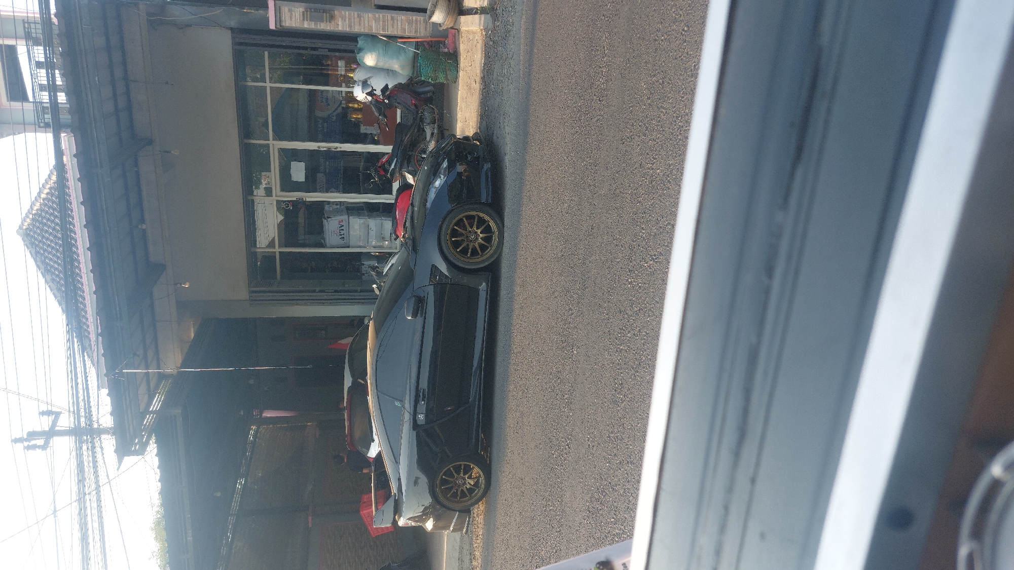 The image depicts a black modified Toyota Celica parked on the side of a street in what appears to be a commercial area of Karas, Indonesia. The car is the central focus, with its sleek design and aftermarket gold rims prominently displayed. Behind the car, a building with a tiled roof and large glass windows suggests it might be a shop or business. Several motorcycles are parked near the building, and some bags are stacked next to them. Power lines and a utility pole are visible in the background, indicating an urban setting. The street is paved with asphalt, and the lighting suggests it is daytime, possibly late afternoon due to the shadows. In the foreground, the bottom of the image is blurred, showing what looks like the edge of a car window or a reflective surface.