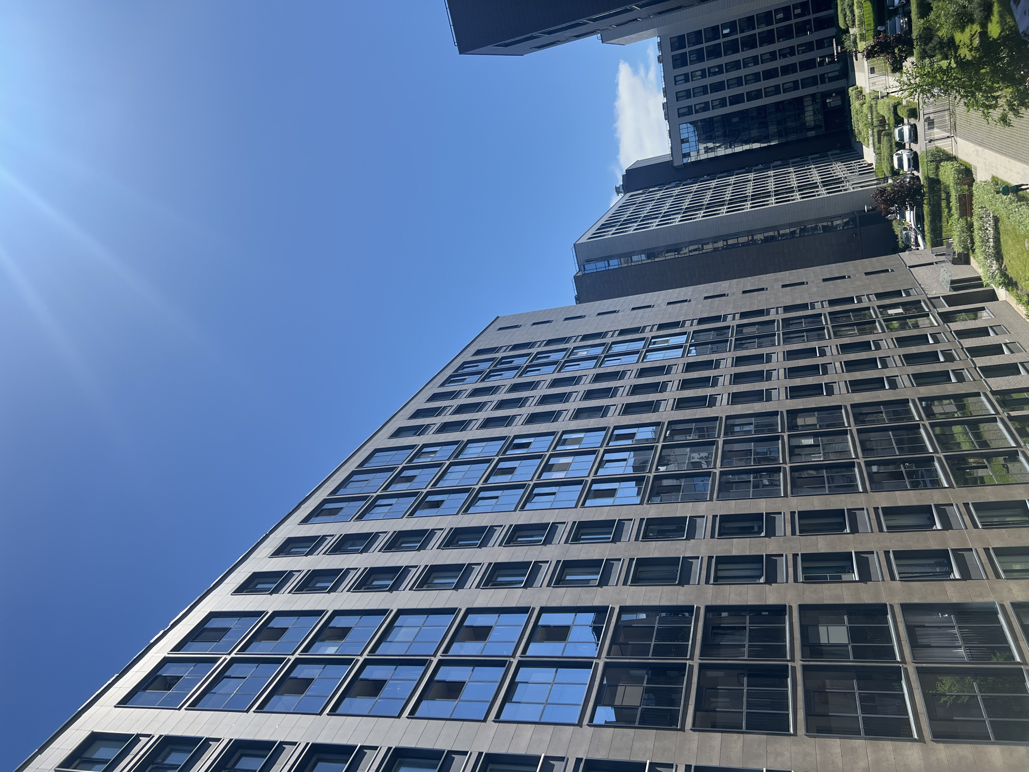 This image depicts a modern residential complex in the city of Saint Petersburg, Russia, viewed from a slightly elevated perspective. The scene is bathed in bright sunlight under a clear, intense blue sky, with a visible lens flare originating from the upper left indicating direct sunlight. A small, wispy white cloud is visible in the upper right sky.
Multiple multi-story apartment buildings with dark, possibly brownish-gray or anthracite facades dominate the frame. The buildings feature numerous uniformly sized, rectangular windows, many of which reflect the vibrant blue sky and some surrounding architecture. The leftmost building extends vertically beyond the frame, showcasing many floors of these reflective windows. Other similar buildings are arranged around a central, landscaped courtyard, forming an urban canyon effect.
The ground level between the buildings consists of meticulously maintained green spaces, including lawns and geometric arrangements of low-lying shrubs. Paved walkways, made of light-colored pavers, traverse these green areas. In the mid-ground, a designated outdoor parking area is visible, where at least three white sedans are parked. A dark metal gate or fence separates a paved access road from the main courtyard. A dark bench is situated on a lawn section. One individual, wearing dark clothing, is seen walking on a paved path in the lower-middle section of the courtyard, moving away from the viewer. The overall environment suggests a contemporary and well-maintained urban living space.