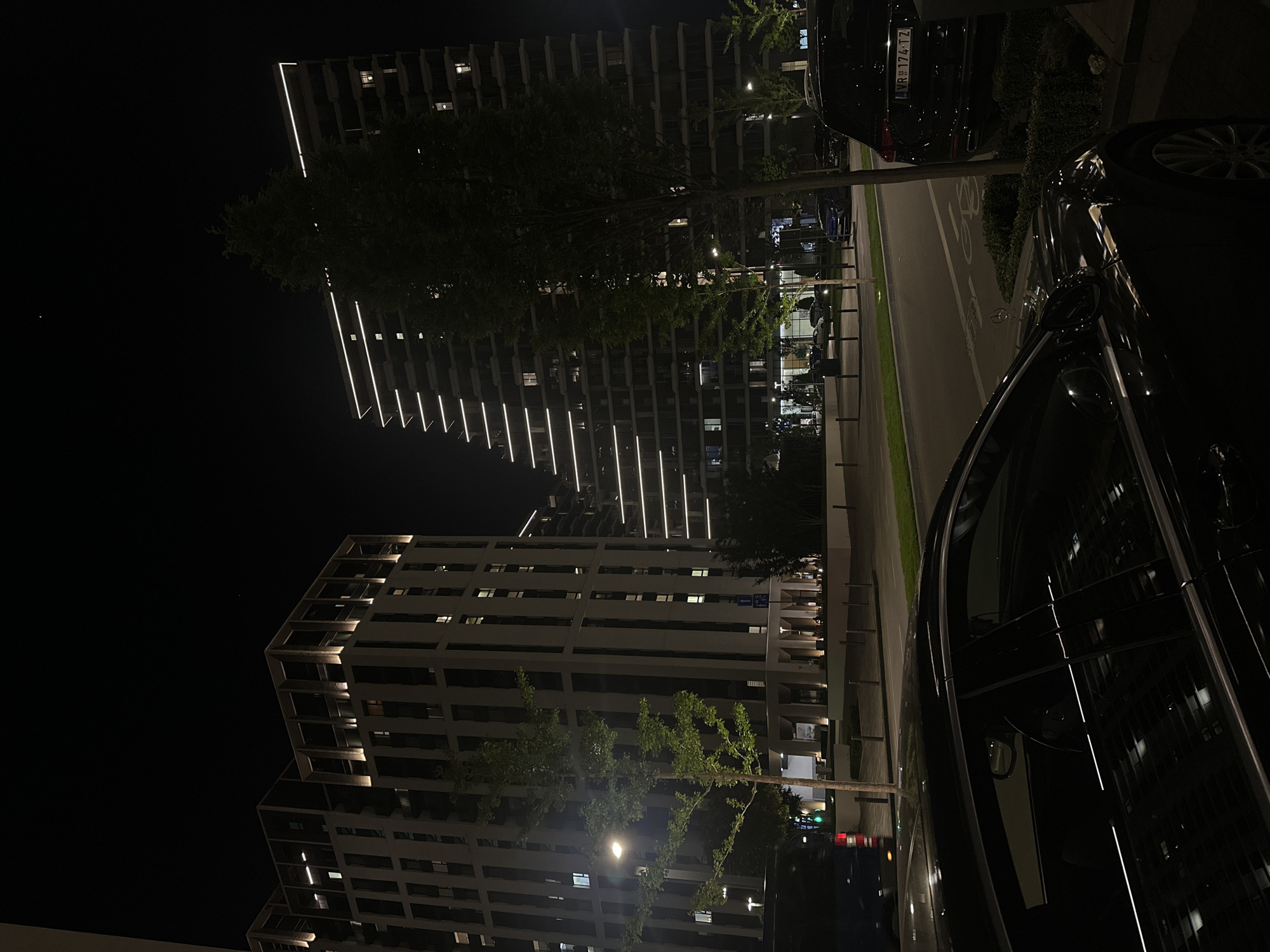 The image depicts a nighttime scene in Belgrade, Serbia, showcasing modern architecture illuminated against the dark sky. Two high-rise buildings are prominent, one featuring a distinctive angular design with illuminated lines ascending diagonally. The other building exhibits a more conventional rectangular structure with lit windows. Several trees partially obscure the building facades. A street runs alongside the buildings, with parked cars lining the edge, including at least one visible with a license plate "MR 174 TZ". The street also includes a bicycle lane. The overall environment is urban and well-lit, typical of a modern city at night.