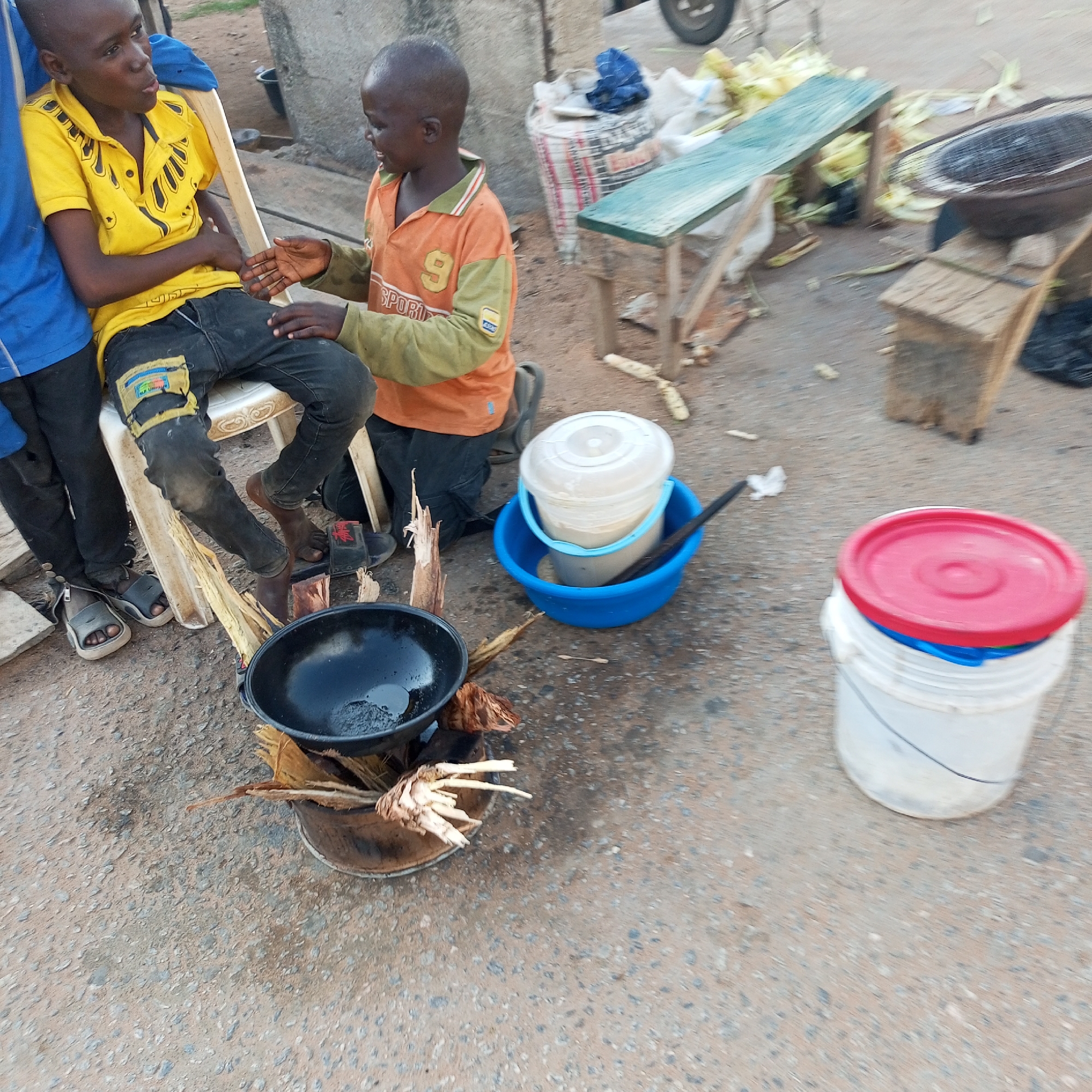 Two young boys are seen outdoors in Jalingo, Nigeria. One boy, wearing a yellow shirt with black designs and dark pants, sits on a white chair. The other boy, in an orange long-sleeved shirt with a yellow collar and a number nine on the front, kneels nearby. They appear to be engaged in a conversation, with the boy in orange gesturing with his hands.
In the foreground, a black pot sits on what appears to be a makeshift fire pit with dried plant material. To the right, a blue bucket holds a white container, and next to that, a white bucket with a red lid is visible. The ground is a mixture of dirt and gravel, typical of outdoor settings in Jalingo. In the background, there are some wooden structures and bags, suggesting a market or roadside vending area. The overall scene depicts a common street-side activity.
