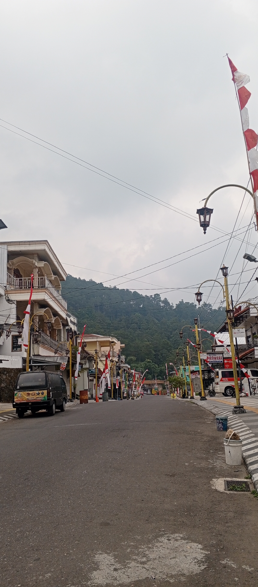 The image depicts a street scene in Sarangan, Indonesia, likely on a cloudy day given the overcast sky. The street is lined with buildings on both sides, adorned with numerous red and white flags, suggesting a national holiday or celebration. The buildings appear to be a mix of commercial establishments and residences, with balconies and signage visible. One building on the left has a sign that appears to say "HOTEL NIRWANA."
Streetlights with a decorative, traditional design are spaced along the street. A dark-colored van is parked on the left side of the road, and further down, a white van with red accents is also visible, along with a motorcycle. The road surface is asphalt, with a striped pedestrian crossing in the foreground.
In the background, a lush, forested hillside rises, indicating the natural landscape surrounding the town. The overall impression is of a quiet, somewhat hilly town decorated for a special occasion. There are no people clearly visible in the image.