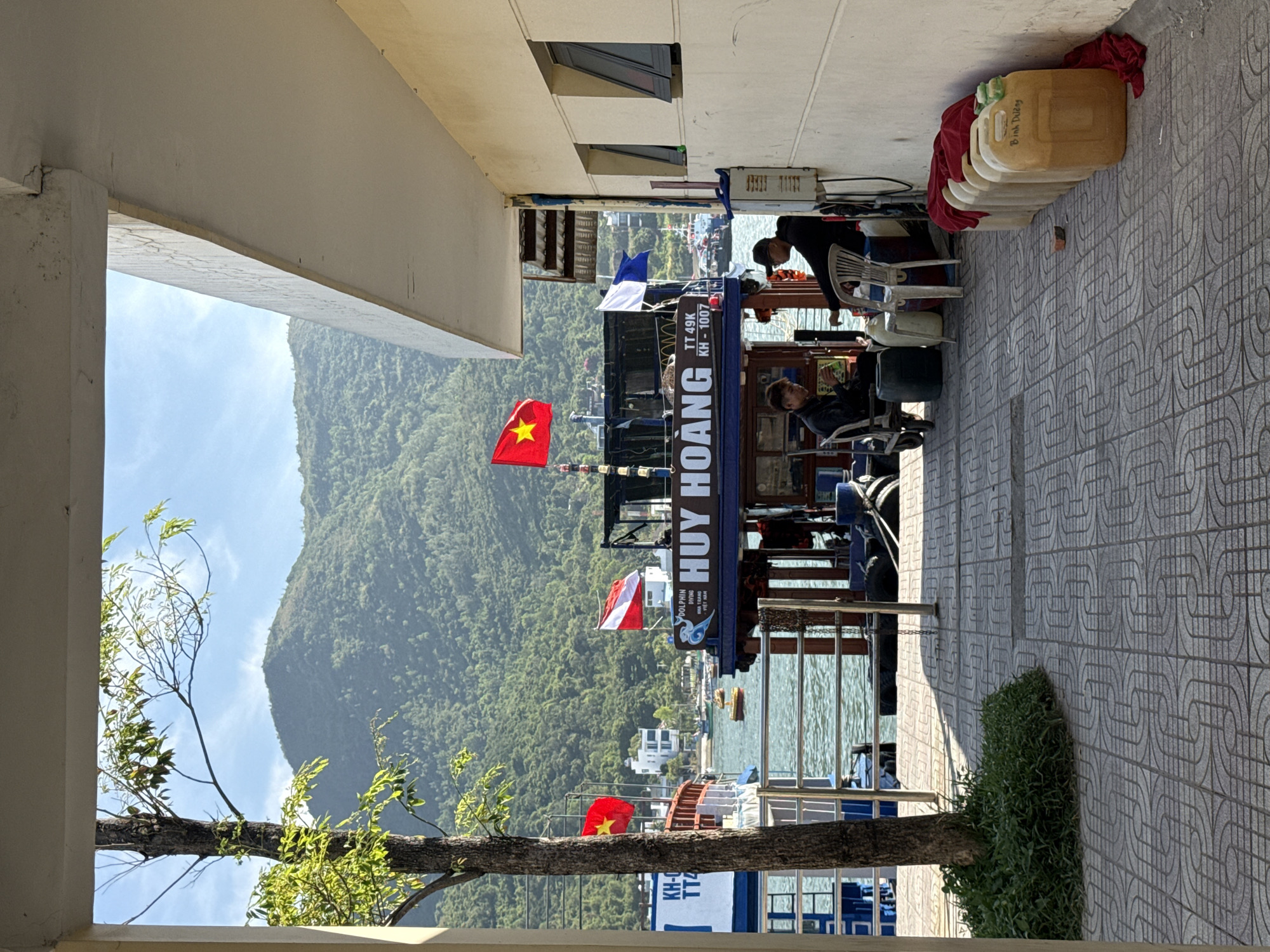 The image, rotated for viewing, depicts a scene in Nha Trang Ward, Vietnam, potentially a coastal area. Framed by a concrete structure, a boat named "HUY HOANG" is prominently displayed, bearing Vietnamese flags. The boat also features the text "DOLPHIN DIVING NHA TRANG VIETNAM" and the registration numbers "TT 49K KH-1007". Two individuals are visible on the boat. One is seated and appears to be using a device, while the other is standing and possibly working. The background reveals a mountain range under a clear sky. The foreground shows a tiled pathway with a patch of vegetation. Containers, chairs, and clothing are stacked near the concrete structure, suggesting an active workspace. The OCR text on the image confirms its location and details of the boat.