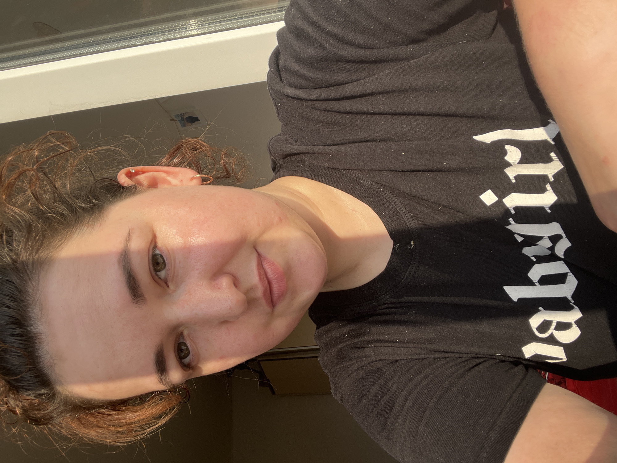 A young woman with brown, curly hair is captured in a close-up, selfie-style photograph. She is wearing a black t-shirt with the white gothic font lettering "babygirl" emblazoned across it. Her gaze is directed towards the camera. The lighting suggests it is either early morning or late afternoon, with warm sunlight casting shadows across her face. A window frame is visible on the right side of the image, hinting at an indoor setting. The backdrop appears to be a domestic environment, with hints of furniture and decor in the background. The image is likely taken within a residential setting in the city of Chelyabinsk, Russia.