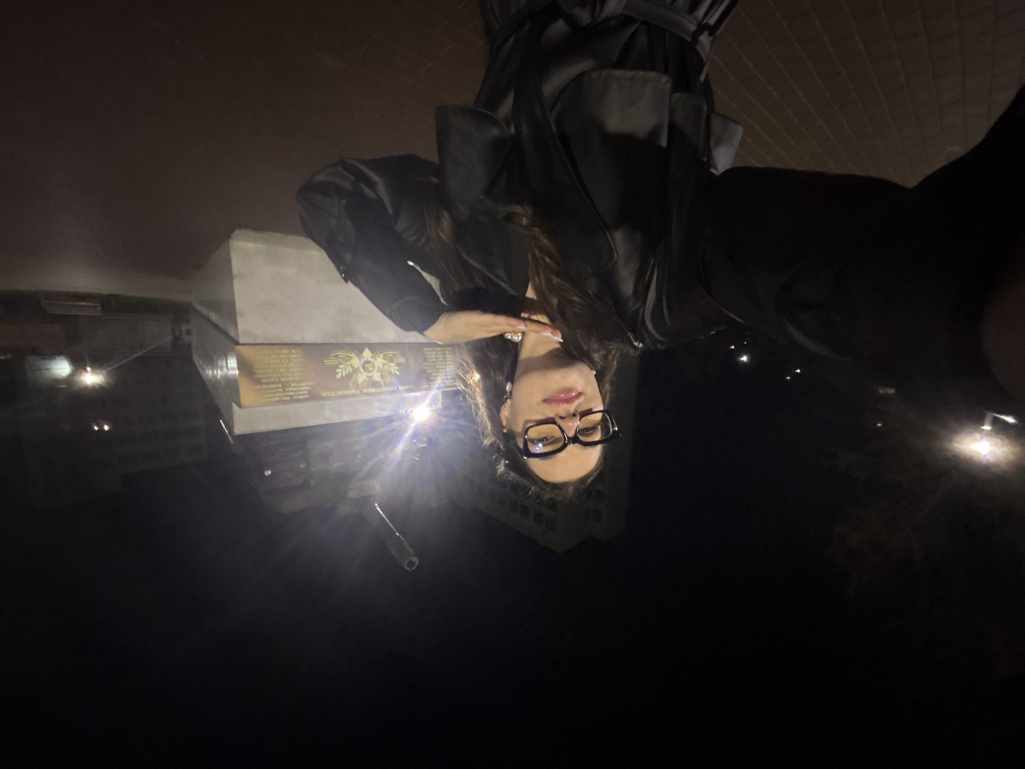 A woman with long dark hair is posing for a photo in Chaikovsky, Russia, at night. She is wearing glasses and a dark coat. Behind her is a monument. Streetlights illuminate the scene. In the background, buildings are dimly visible.
