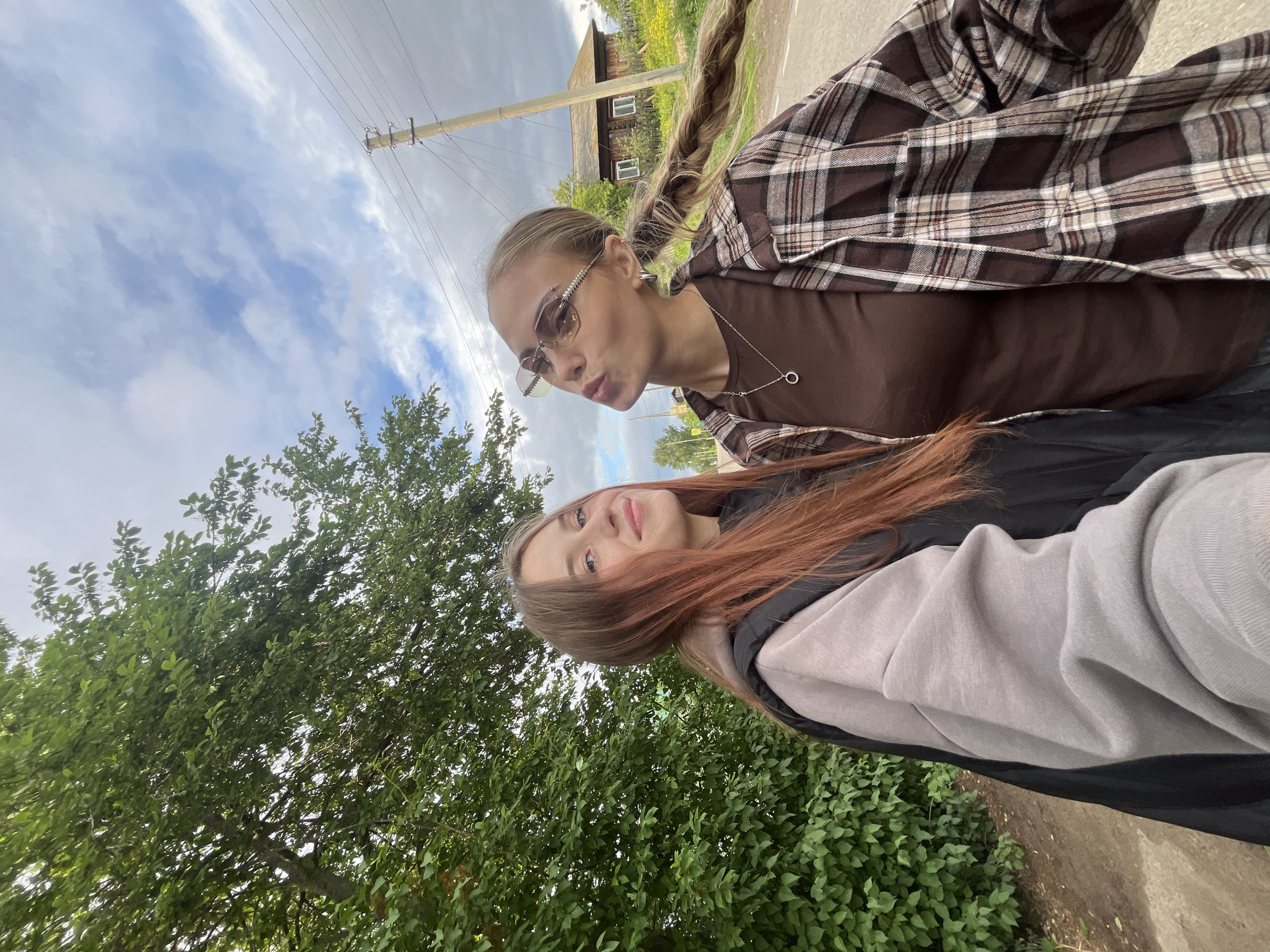 Two young women are captured in a daytime outdoor photo, likely a selfie, in a rural or semi-rural setting identified as Фоки, Russia. The image is taken from a low angle, looking upwards towards the subjects and the sky.
The woman in the foreground on the left (when the image is rotated correctly) has long blonde hair styled in a thick braid, which falls over her right shoulder. She is wearing stylish, light-tinted sunglasses with decorative elements on the frames. Her expression is playful, with her lips puckered in a "duck face." She is dressed in a dark brown top beneath a brown, white, and black plaid overshirt. A delicate silver chain with a circular pendant hangs around her neck.
Partially visible next to her is another young woman with long reddish-brown hair. She has a subtle smile and appears to be wearing a light grey hooded sweatshirt under a black jacket.
The background features a cloudy sky, with patches of blue visible between the white and grey clouds. Lush green trees and bushes frame the lower and left portions of the image. In the mid-ground, a wooden utility pole with wires is visible, extending towards a distant, simple wooden house with small white-framed windows, surrounded by green and yellow foliage. The overall atmosphere suggests a casual moment outdoors on a cloudy day. There is no visible text in the image.