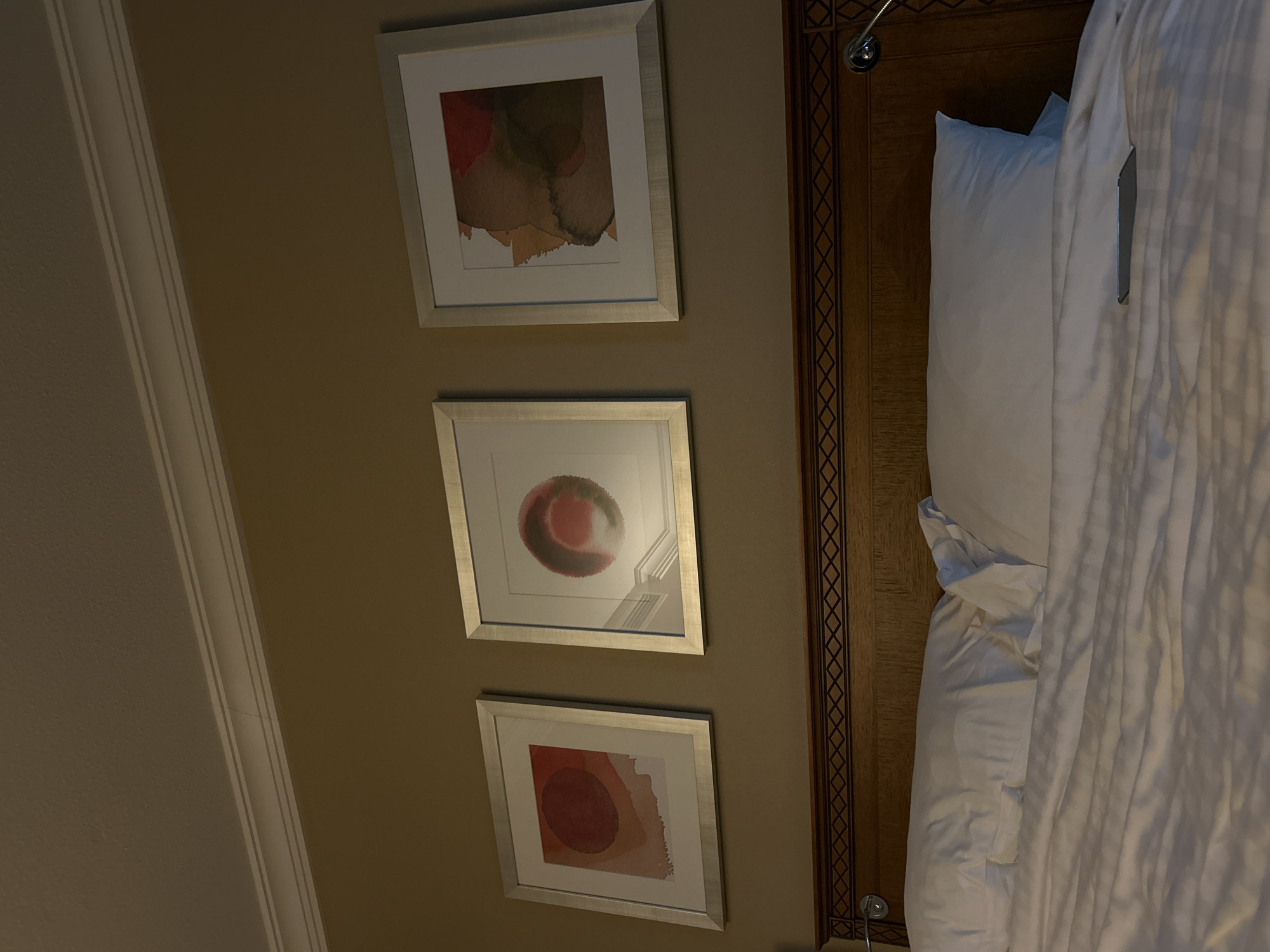 The image shows a portion of a room interior with a bed and wall decorations. Three framed artworks, abstract in style, are aligned vertically on a beige wall. Each artwork has a silver frame. The top artwork features brown and red hues, the middle artwork shows a red circular form, and the bottom artwork also displays a red circular form with additional colors. Adjacent to the artwork is a headboard with decorative patterns, and part of a bed with white bedding. It is not possible to ascertain the exact location or city from the available details.