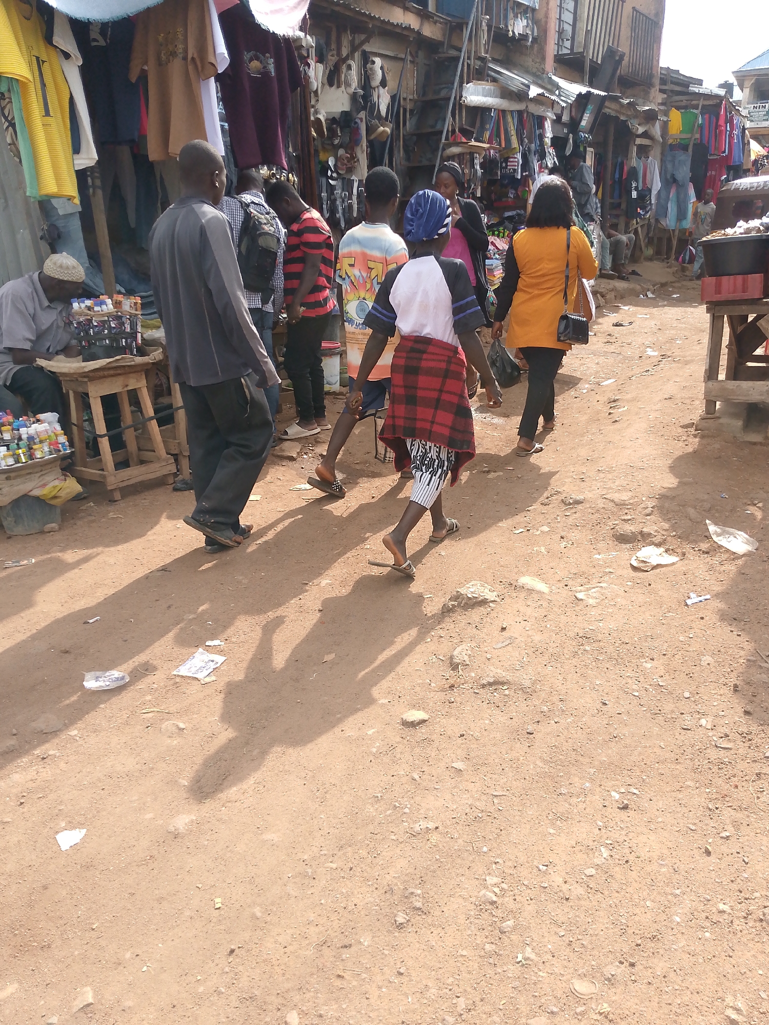 This image captures a bustling outdoor market scene in Bukuru, Nigeria. The ground is a dusty, reddish-brown earth, scattered with small pieces of debris. Several people are walking through the market, some heading away from the camera.
In the foreground, a young person is walking away, wearing a white and black t-shirt with blue accents, and a red and black plaid skirt wrapped around their waist. Below the skirt, striped leggings are visible. To their left, a man in a grey long-sleeved shirt and dark pants walks away, also in flip-flops. Behind them, a person in a red and black striped shirt and dark pants walks. Further back, a person in an orange top and black pants carries a black handbag.
The background shows stalls filled with clothing. One stall has an array of brightly colored t-shirts hanging, including yellow, brown, and maroon. Another stall displays more clothing and what appear to be shoes. The structures of the stalls are rustic, suggesting a temporary or open-air market. A man wearing a cap and a grey shirt sits behind a small wooden table displaying bottles of what looks like spices or small goods, suggesting a vendor. The sunlight indicates it is daytime, likely mid-morning to afternoon, with clear, bright conditions casting distinct shadows on the ground. The overall atmosphere appears active and busy, typical of a local market.