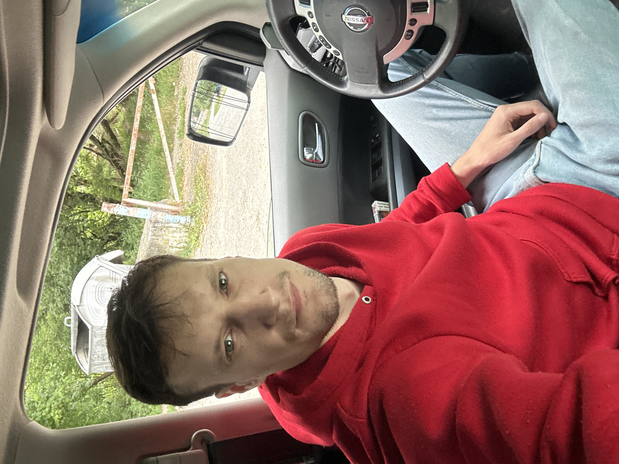 In Makiv, Ukraine, a man is pictured inside a vehicle, likely a Nissan given the logo on the steering wheel. He is wearing a red hooded sweatshirt and light blue jeans. The interior of the car is visible, including the dashboard, steering wheel, and door panel. Outside the car, through the windshield and side window, a rustic gate and a large, metal dumpster can be seen amidst lush greenery, suggesting a rural or semi-rural setting. The man is looking directly at the camera.