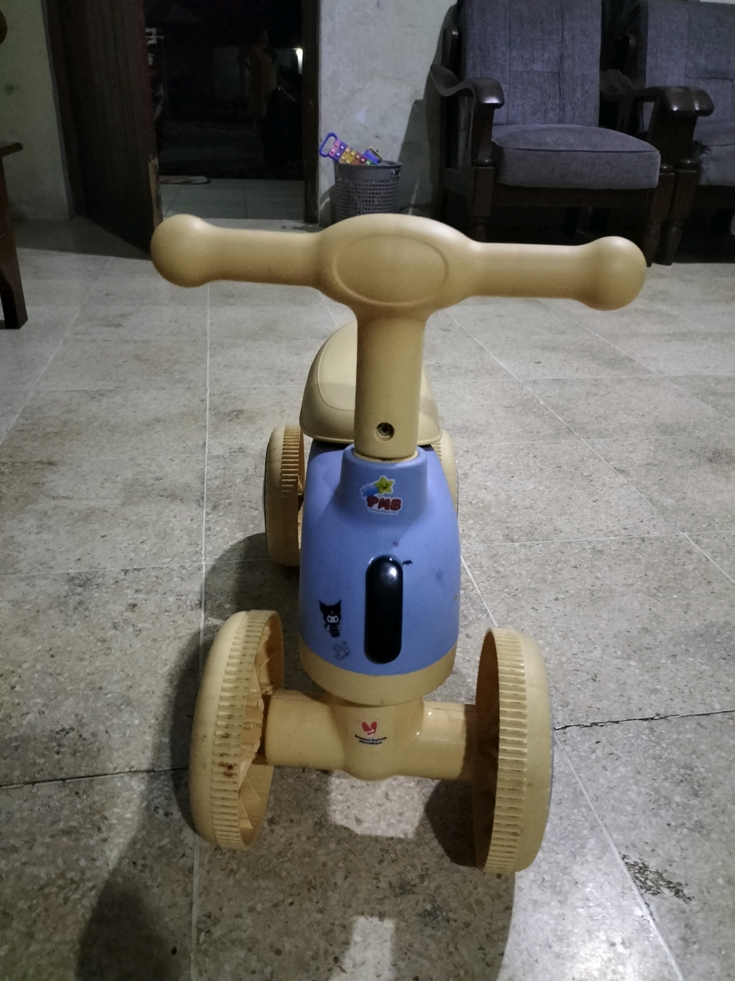 A low-angle, close-up, full-shot image shows a small, plastic, ride-on toy for a toddler. The toy is primarily yellow and blue, with a yellow handlebar, yellow seat, and a blue body. It has four yellow wheels, two in the front and two in the back, with ridged edges. A black oval-shaped piece is visible on the blue body. There are some cartoon stickers on the blue body, including a black cat-like character. The toy is on a floor made of gray tiles. In the background, a doorway leads to a dark room, and two upholstered chairs are visible. The lighting is dim, casting shadows on the floor. The scene appears to be an indoor residential setting in Tasikmadu, Indonesia.