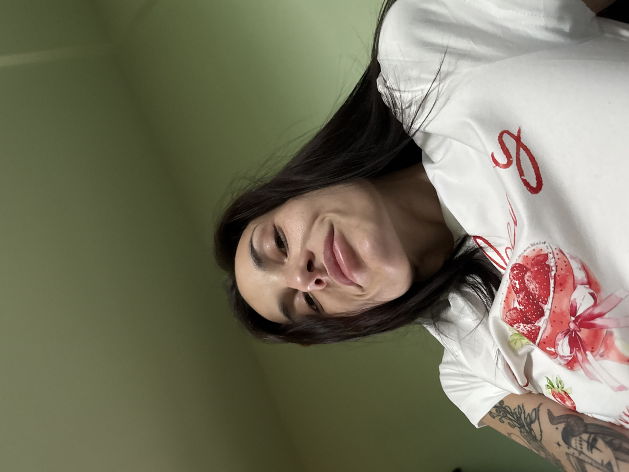 A low-angle, close-up photograph depicts a female subject indoors against a plain olive-green wall. The woman has long, dark, straight hair, an oval-shaped face with prominent cheekbones, visible freckles across her nose and cheeks, and full, slightly parted lips with a glossy finish. She is looking upwards, directly at the camera, with a subtle smile. She wears a white short-sleeved t-shirt featuring a central graphic of a red strawberry dessert in a white dish with a red bow, alongside partially visible red cursive text. Dark, intricate tattoos cover her left arm, including what appears to be a stylized head or figure on the bicep area. No specific location within Pleven, Bulgaria, is identifiable from the visual content of the image.