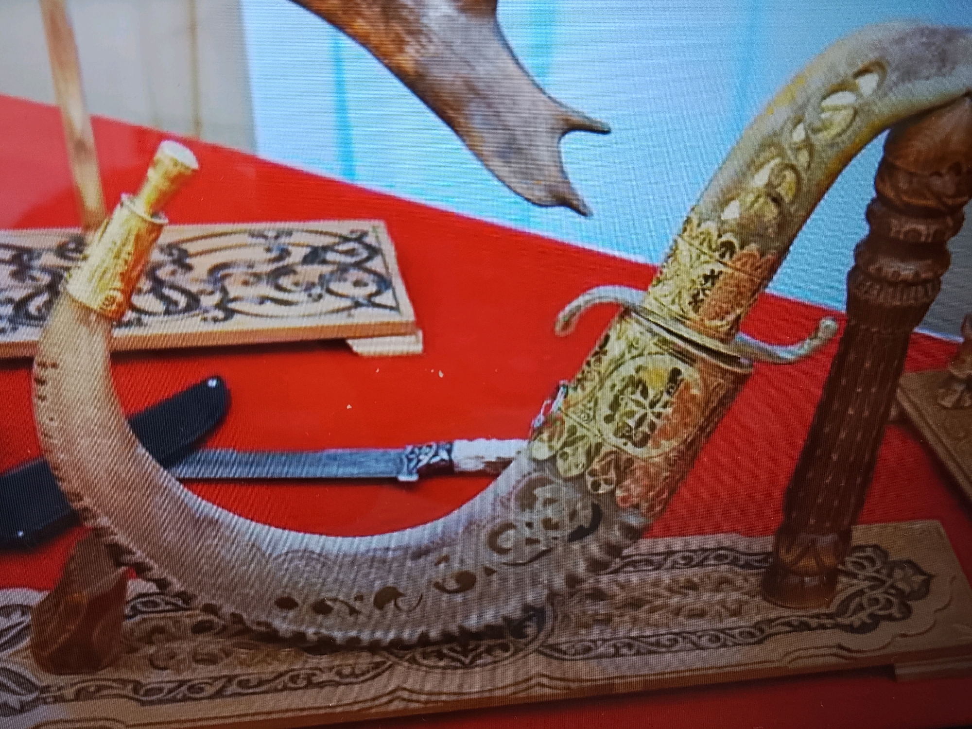 A curved, ornate dagger with a golden hilt and scabbard rests on a decorative wooden stand. The dagger's blade is visible, showing intricate carvings. Another smaller knife with a black handle is partially visible behind the dagger. In the background, a piece of a large antler is also present. The scene is set against a red surface, possibly a tablecloth, with a light blue backdrop. This image was taken in Doha, Qatar.