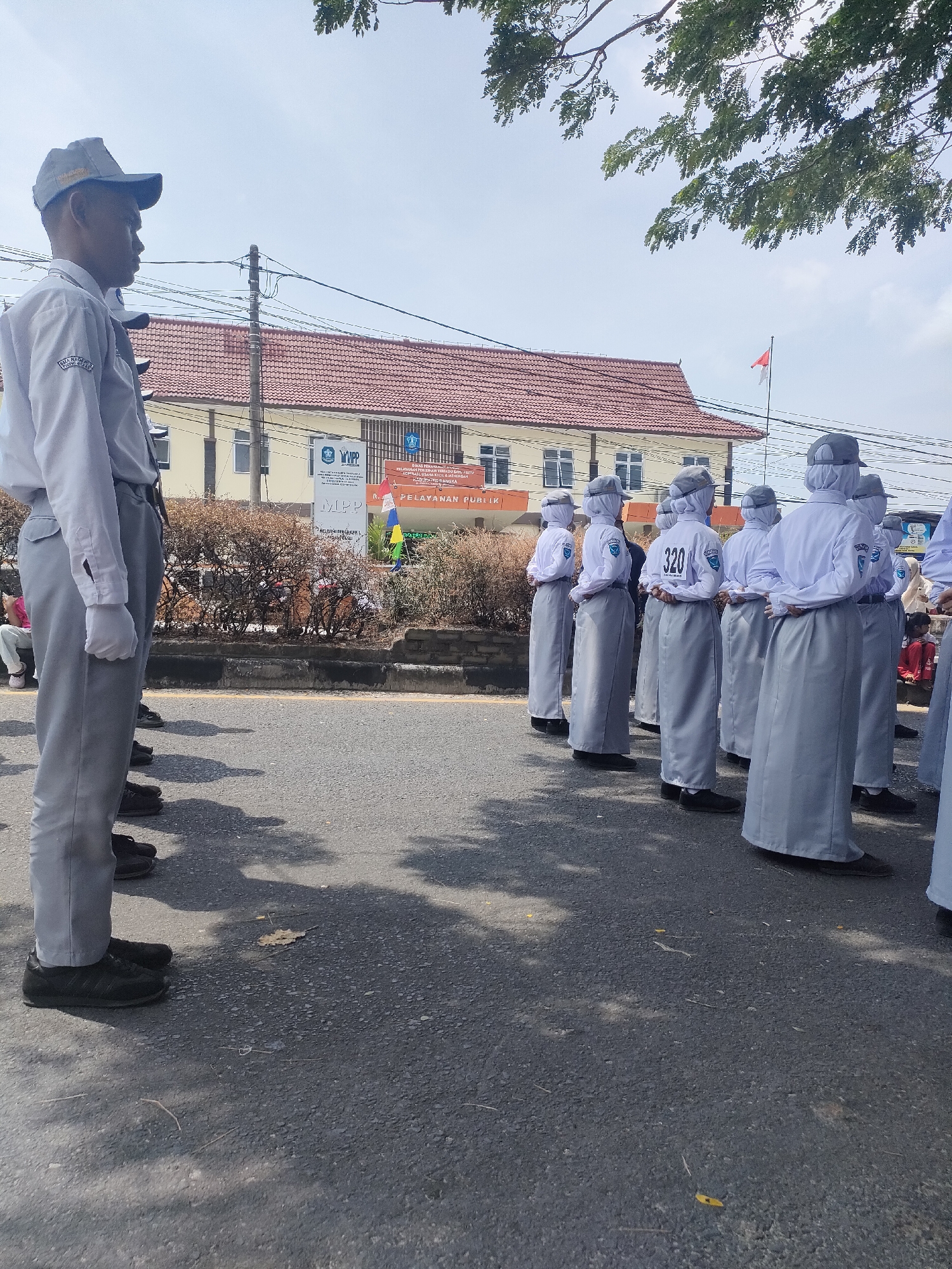 In Sungailiat, Indonesia, a group of students, likely high school age, stand in formation on an asphalt road. They are dressed in identical uniforms consisting of white long-sleeved shirts with blue emblems on the sleeves, grey headscarves or caps, and long grey skirts or pants. Some students have numbers visible on their backs, such as "320." They are facing away from the camera, looking towards a building in the background. This building appears to be a public service center, with signage that includes "PELAYANAN PUBLIK" (Public Service) and "MPP". The building has a red tiled roof and is bordered by dry shrubbery. A red and white Indonesian flag flies from a flagpole in front of the building. The sky is clear and blue, and shadows from trees are cast on the road. The overall scene suggests an outdoor event or ceremony.