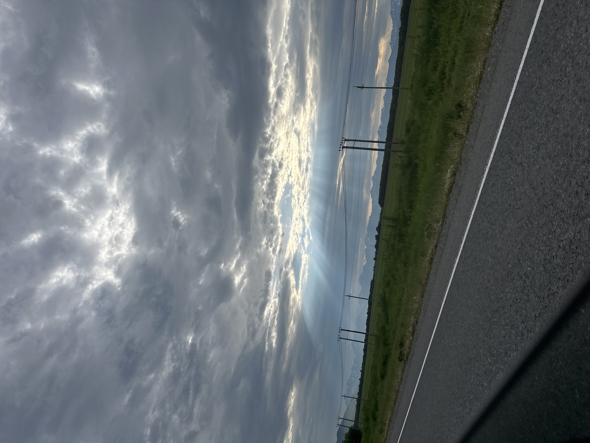 This is an outdoor, eye-level, medium shot photograph of a rural landscape with a stormy sky. The image is taken from a moving vehicle, as evidenced by the shadow of the vehicle falling across the lower left portion of the frame and the angle of the road. The road, composed of asphalt, runs diagonally from the bottom left to the middle right of the frame. A white line marks the edge of the lane closest to the viewer.
To the right of the road is a grassy field with scattered small white flowers. In the distance, a line of trees can be seen bordering the field. Further back, a range of snow-capped mountains stretches across the horizon under a dramatic sky. The sky is filled with heavy, dark gray clouds, with breaks allowing bright sunlight to stream through in visible rays. These sunbeams create shafts of light that illuminate parts of the landscape and the sky. Several utility poles with power lines are visible in the middle ground, spaced out across the field. The overall atmosphere suggests an impending or recently passed storm. There are no people or animals visible in the frame. The specific location of Кеткино, Russia, is not visually determinable from the provided image.