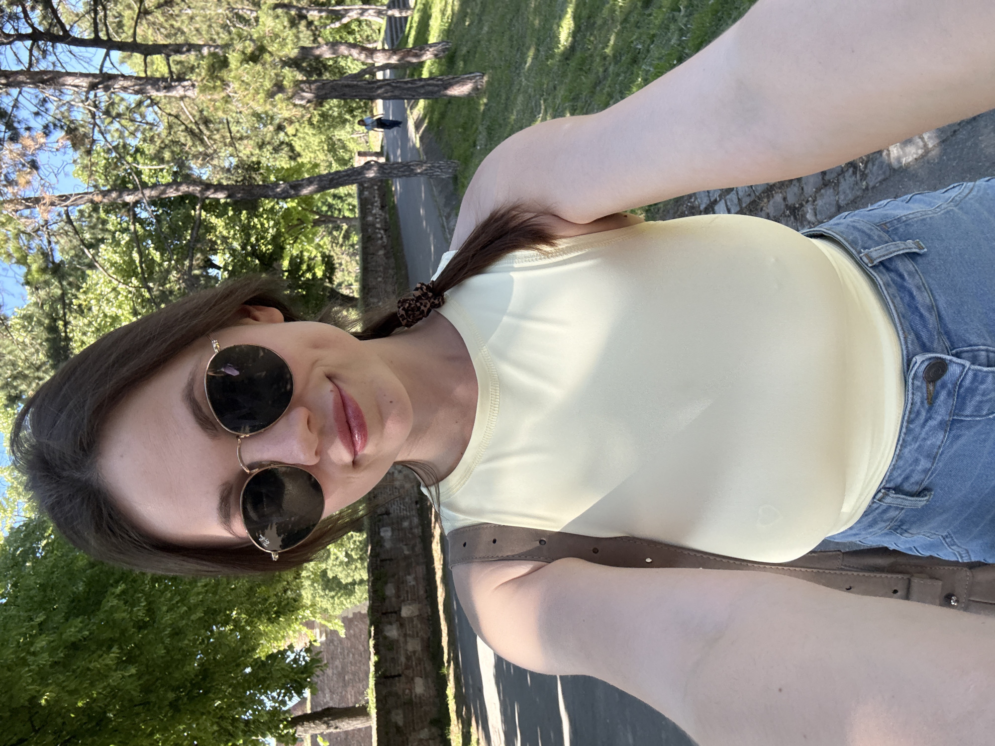 A young woman is taking a selfie in a park, likely in Belgrade, Serbia. She is wearing sunglasses, a yellow top, and jeans, with a brown belt. The background includes trees, a paved path, and a stone wall. Another person is visible walking further along the path. The light suggests it is a sunny day.