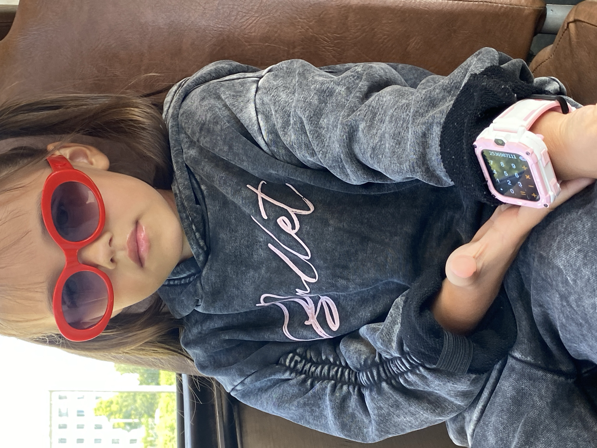 A young girl with brown hair in pigtails, wearing red oval sunglasses and a dark grey acid-wash hoodie with pink cursive writing, sits on a brown seat. A small pink and white digital watch is visible on her left wrist. The background includes a window with a view of a building and trees, suggesting an urban environment, potentially in Chaikovsky, Russia. The lighting indicates it is daytime.