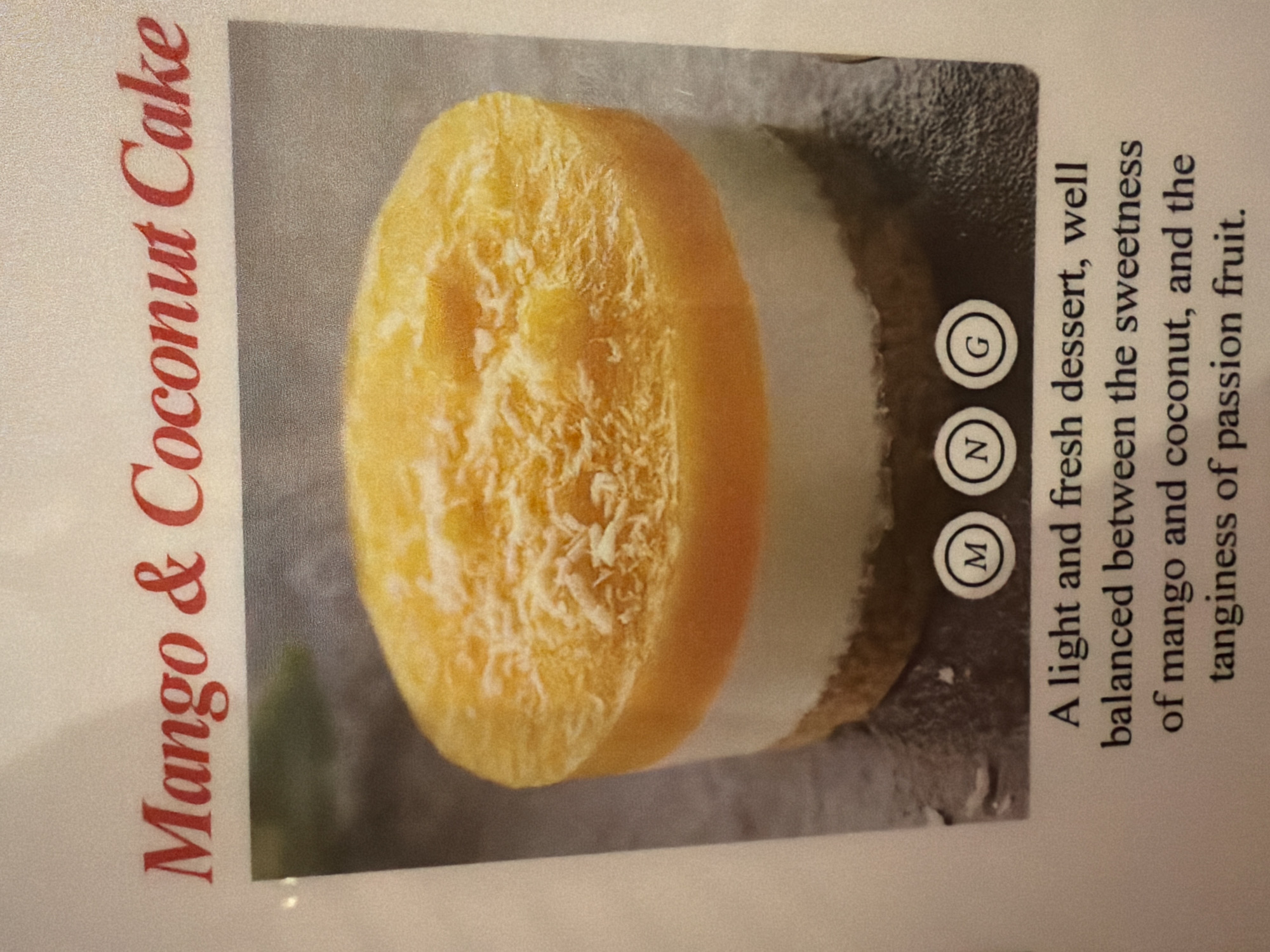 The image is a close-up shot of a menu item: Mango & Coconut Cake. A round, multi-layered cake is prominently displayed. The top layer is a bright yellow mango color, textured with what appears to be shredded coconut. Beneath the mango layer is a white layer, and below that is a darker, possibly coconut-based layer. To the right of the cake are three circled letters: "M," "N," and "G." Next to the cake is a description of the dessert: "A light and fresh dessert, well balanced between the sweetness of mango and coconut, and the tanginess of passion fruit." The text is in black. The dessert and text are set against a gray background. The words "Mango & Coconut Cake" are printed in red on the left side of the image. There is no evidence of the city of London, United Kingdom in the image.