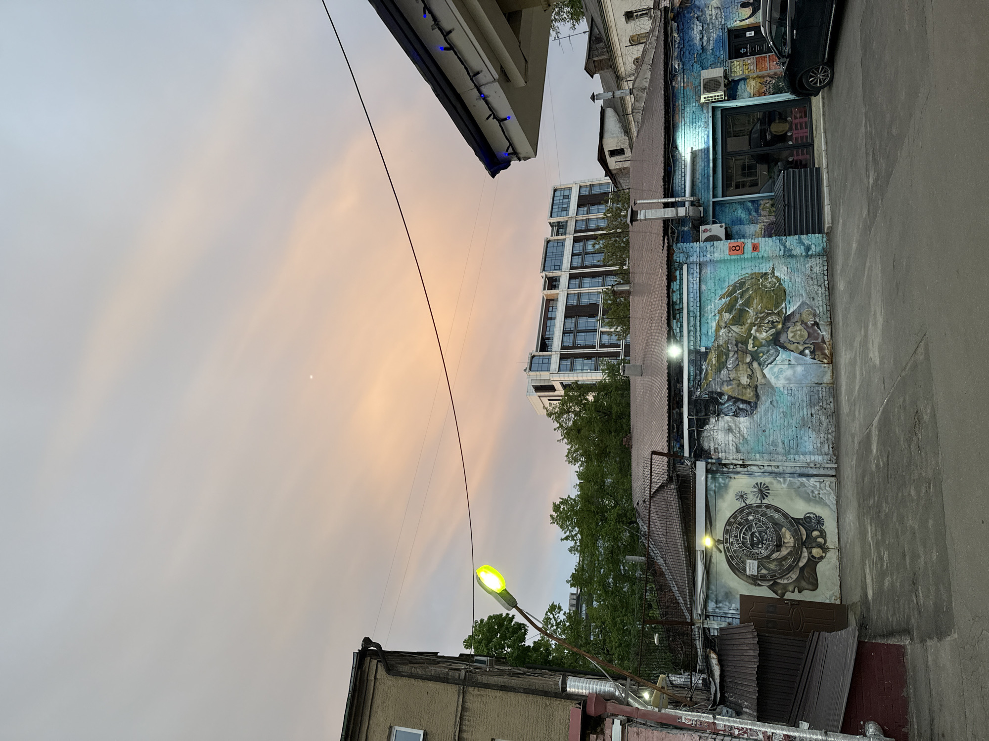 The image presents an urban scene likely in Moscow, Russia. The sky is overcast with a pink-orange hue. A street lamp emits a green light. Buildings of varying heights and architectural styles surround the street. One building exhibits a mural. A car is partially visible in front of one of the buildings. The scene appears to be taken from an elevated position.