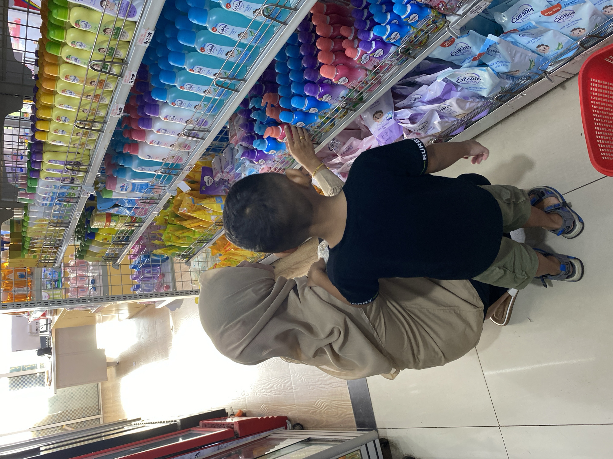 In the city of Lampisang, Indonesia, two individuals, a woman wearing a tan hijab and a young boy in a black t-shirt and khaki shorts, are browsing a brightly lit aisle of a retail store, likely a minimarket or convenience store. They are standing in front of shelves stocked with various personal care products, including bottles of lotion or baby wash in assorted colors and packaging, as well as individually wrapped wet wipes. The woman is slightly crouched down, and the boy is reaching towards a shelf. The floor is tiled in a light color, and sunlight streams in from an unseen source, casting shadows. To the left, a red shopping basket is visible. The environment appears clean and organized, typical of a retail establishment.