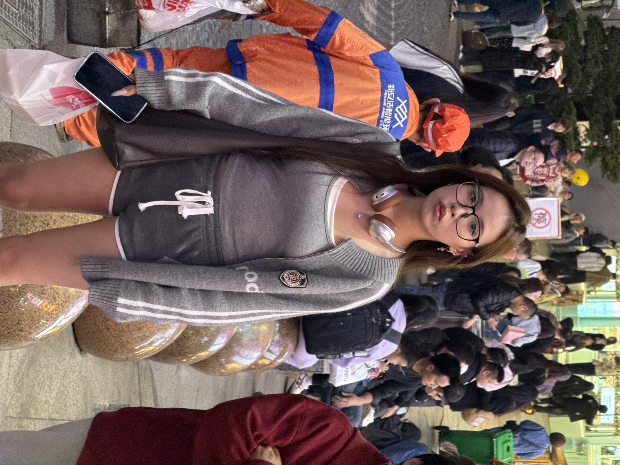 The scene depicts a bustling urban pedestrian area in Liangjiang, China. In the foreground, a young woman stands centrally, facing forward with a neutral expression. She has long brown hair, wears black-rimmed glasses, and has white and silver wireless headphones draped around her neck. Her attire consists of a grey tank top, a grey zippered cardigan with white stripes on the sleeves and a shield emblem on the left chest, and dark grey shorts with white trim and a white drawstring. She holds a black smartphone and a white plastic bag in her left hand, which hangs at her side.
Immediately to the woman's left and slightly behind her, a person in an orange and blue utility uniform is partially visible. The uniform features Chinese text, "新安洁智慧环卫," which translates to "NEWANGE AMBIENT INTELLIGENT." To the woman's right, a series of large, translucent, sphere-shaped objects filled with a granular material are arranged on the paved ground, serving as seating or decorative barriers.
The background is densely populated with numerous individuals of various ages and genders, engaged in walking, standing, and sitting. Several people are observed using mobile phones. Distant green trees are visible, along with illuminated interiors of commercial buildings. A green wheeled trash can is present on the far right side of the background. A white sign with a red circular symbol containing Chinese characters is partially visible in the mid-ground. The overall environment suggests an active public space, possibly a shopping district or plaza.