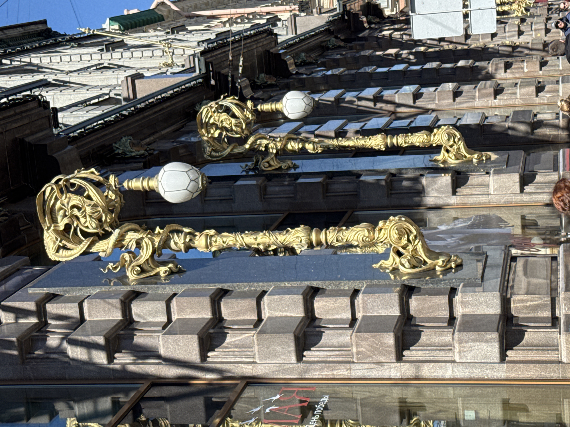 The image shows a close-up view of the facade of the Eliseyev Emporium building in Saint Petersburg, Russia. Two decorative, ornate, gold-colored lamp fixtures protrude from a dark granite-clad portion of the building. Each lamp consists of a fancifully sculpted base, a decorative arm, and a white, globe-shaped lamp. The granite facade is comprised of repetitive, rectilinear blocks. The building's architecture features multiple receding tiers. Parts of the upper levels of the building are visible, as well as reflections in the glass of the lower level. A partial view of a red sign with text in Russian is also visible in the glass reflection. Parts of people are visible at the right side of the image, near the top. The sky is visible at the top. The image appears to be taken during daylight hours.