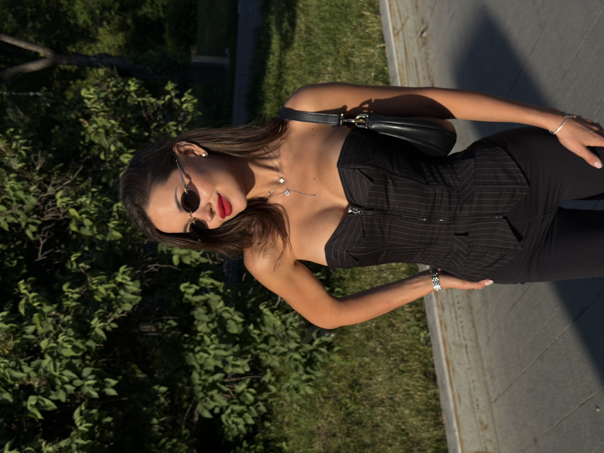The scene is set in an outdoor urban environment, specified as Moscow, Russia, under bright daylight conditions with direct sunlight. A woman, estimated to be in her 20s or 30s, is positioned slightly off-center to the right, looking towards the viewer's right with a subtle smile. She has medium-brown hair, styled straight and falling over her shoulders. She wears small, oval-shaped dark sunglasses with thin gold frames and bright red lipstick. Her accessories include a small silver stud earring in her right ear, a delicate silver Y-shaped necklace with two four-leaf clover-like charms, a silver-toned watch on her right wrist, and a sparkling silver bracelet on her left wrist. She is dressed in a black strapless corset-style top with faint vertical pinstripes and a silver zipper running down the front. This is paired with fitted black pants. A black shoulder bag with a gold-toned clasp is visible on her right shoulder. Her left hand is placed on her hip, displaying light pink manicured nails. The background consists of dense green foliage, including trees and bushes, and a patch of green grass. In the foreground and mid-ground, a gray concrete or paved walkway with visible seams is present, separated from the grass by a low, weathered concrete curb. Distinct shadows are cast on the pavement due to the intense sunlight.