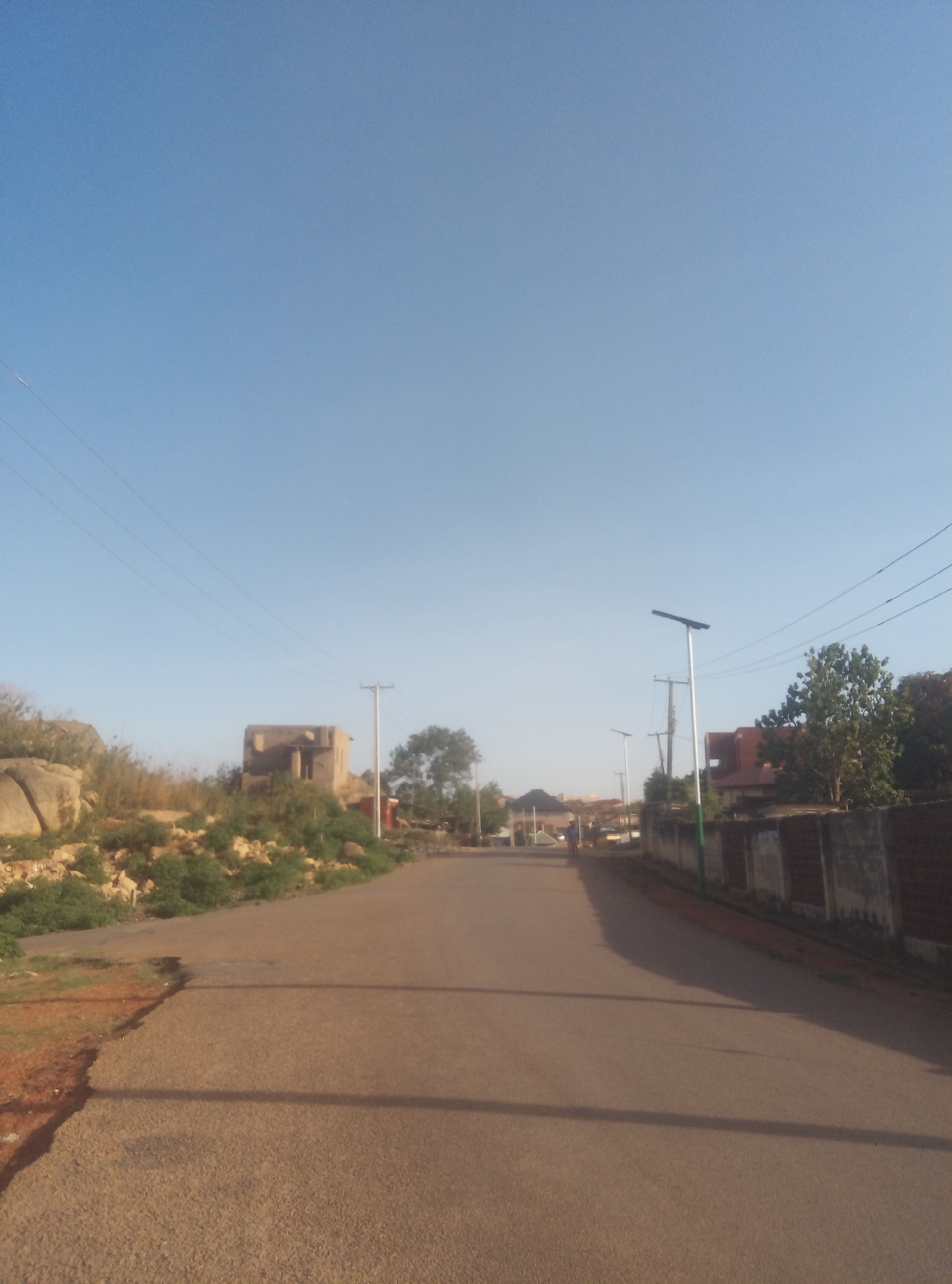The image shows a paved road in a residential area. The sky is clear and blue. On the left side of the road, there's a rocky area with vegetation and a small, old building. Power lines are visible above. On the right side, there's a wall and some trees. In the distance, there are houses and streetlights. A person is walking away from the camera. The location appears to be a quiet street in an urban area.