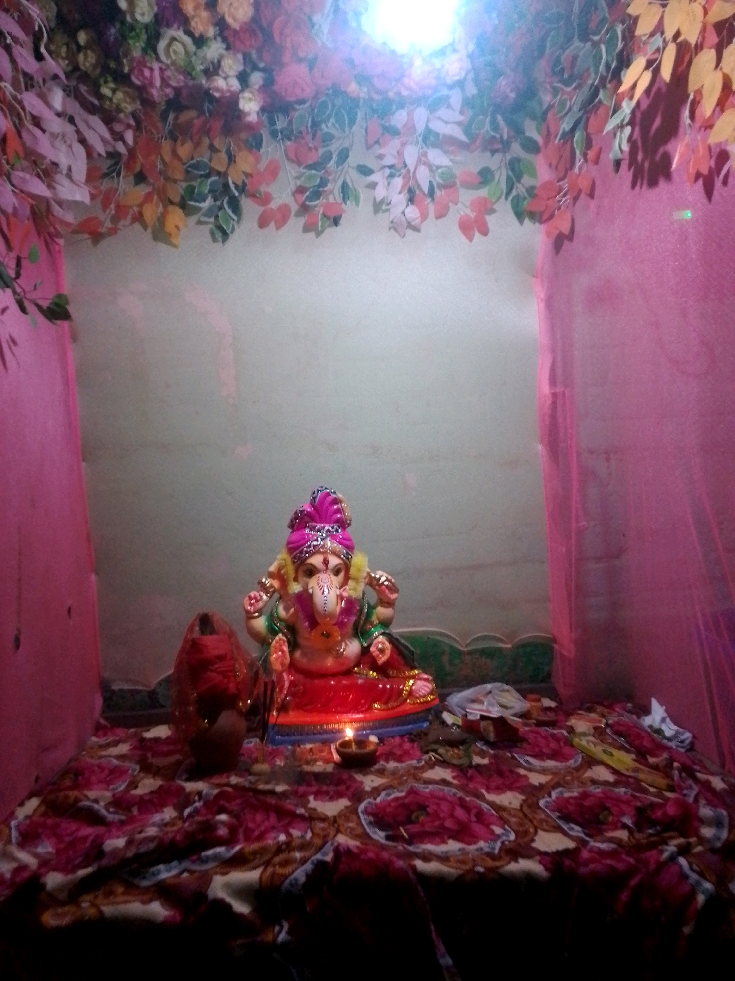 The image depicts an indoor shrine, likely in Siliguri, India, dedicated to the Hindu deity Ganesha. A brightly colored idol of Ganesha, adorned with a pink and gold turban, is centrally placed on a raised platform covered with a patterned cloth. To the left of the idol, a red, conical object, possibly a pot or a decorative item, is visible. The shrine is decorated with artificial flowers and leaves in shades of pink, orange, and yellow, creating an overhead canopy. Pink sheer fabric drapes the sides of the shrine. In the foreground, a patterned cloth covers the surface, displaying floral motifs. A lit oil lamp (diya) and other small offerings are present on the platform. The overall lighting suggests it is either nighttime or a dimly lit indoor space, with a bright light source casting shadows.