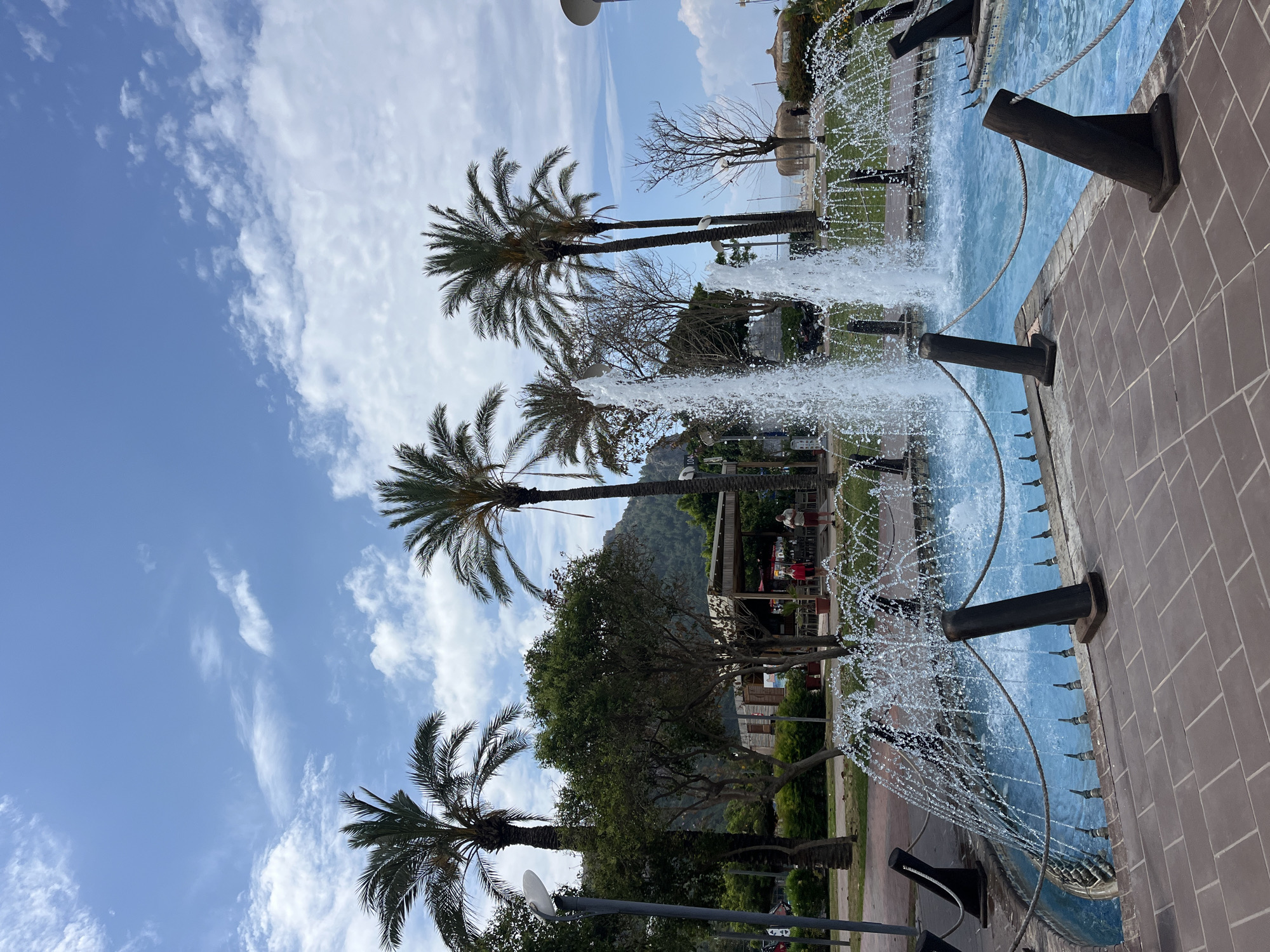 The photograph depicts a landscape featuring palm trees, a fountain, and other urban elements, likely taken in Alanya, Turkey. The sky is partly cloudy, visible between the palm trees. The fountain has several water jets spraying upwards and outwards. The edge of the fountain is constructed from brown brick. Dark, cannon-like structures protrude from the fountain's side. A rope is tied to them. A mountain is visible in the background. A structure resembling a restaurant or covered seating area with red-clad individuals is present in the mid-ground. There are streetlights, other trees, and greenery in the foreground.