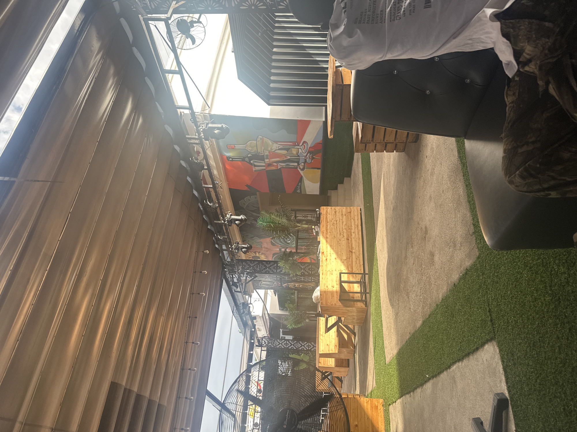 This media file captures an outdoor seating area, possibly a restaurant or bar, with a casual and modern aesthetic. The scene is bathed in bright sunlight, suggesting it's daytime. The setting features a combination of concrete flooring and artificial turf, creating distinct zones. Several wooden benches and tables are arranged to accommodate patrons.
Dominating the upper portion of the image is a large, corrugated metal awning or wall, casting striped shadows across the area. Above this, a ceiling fan is visible, hinting at efforts to keep the space cool. In the background, a colorful mural depicting bottles and glasses adds an artistic touch to the environment. Various metal railings and decorative screens are also present, contributing to the overall design.
A person is partially visible in the foreground, seated on a dark-upholstered chair, with their upper body and arm in view. They are wearing a white t-shirt with text printed on it. The text includes phrases like "SHAME," "REGRET," "PAST MISTA," "UNFORGIVE," "HURT," and "ANGER," followed by "Subtotal," "GRAND T," "FOR THE," and "FIRER GE." A large industrial fan is positioned in the lower part of the frame, suggesting a warm climate. The overall impression is a relaxed outdoor gathering space, likely in a warm climate given the fans and outdoor setup.