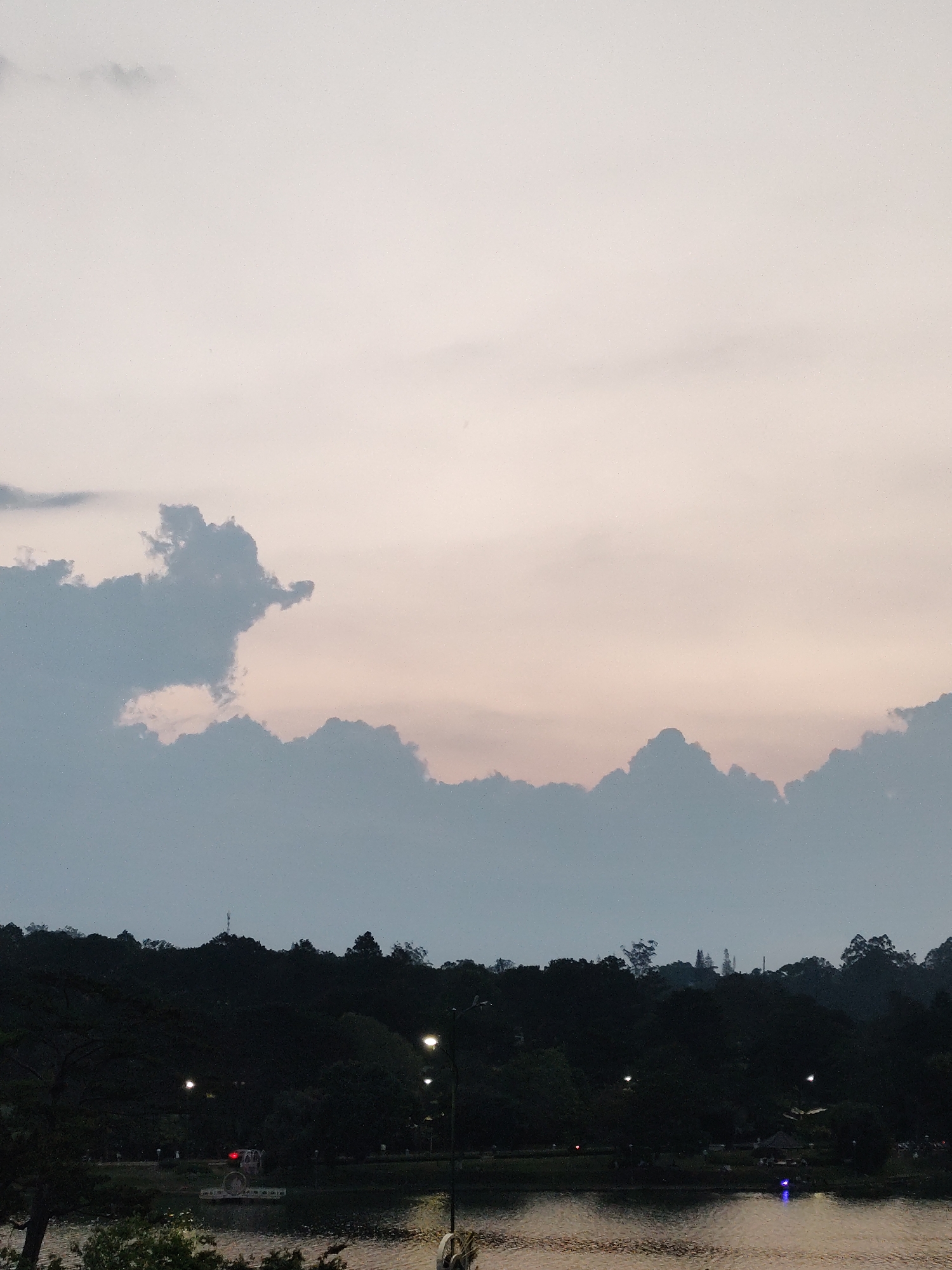 The image shows a twilight scene at Xuan Huong Lake in Da Lat, Vietnam. The sky is light pink and grey with cumulus clouds. A dark green tree line sits between the sky and the lake. Streetlights and possibly a paddle boat are visible near the water’s edge.