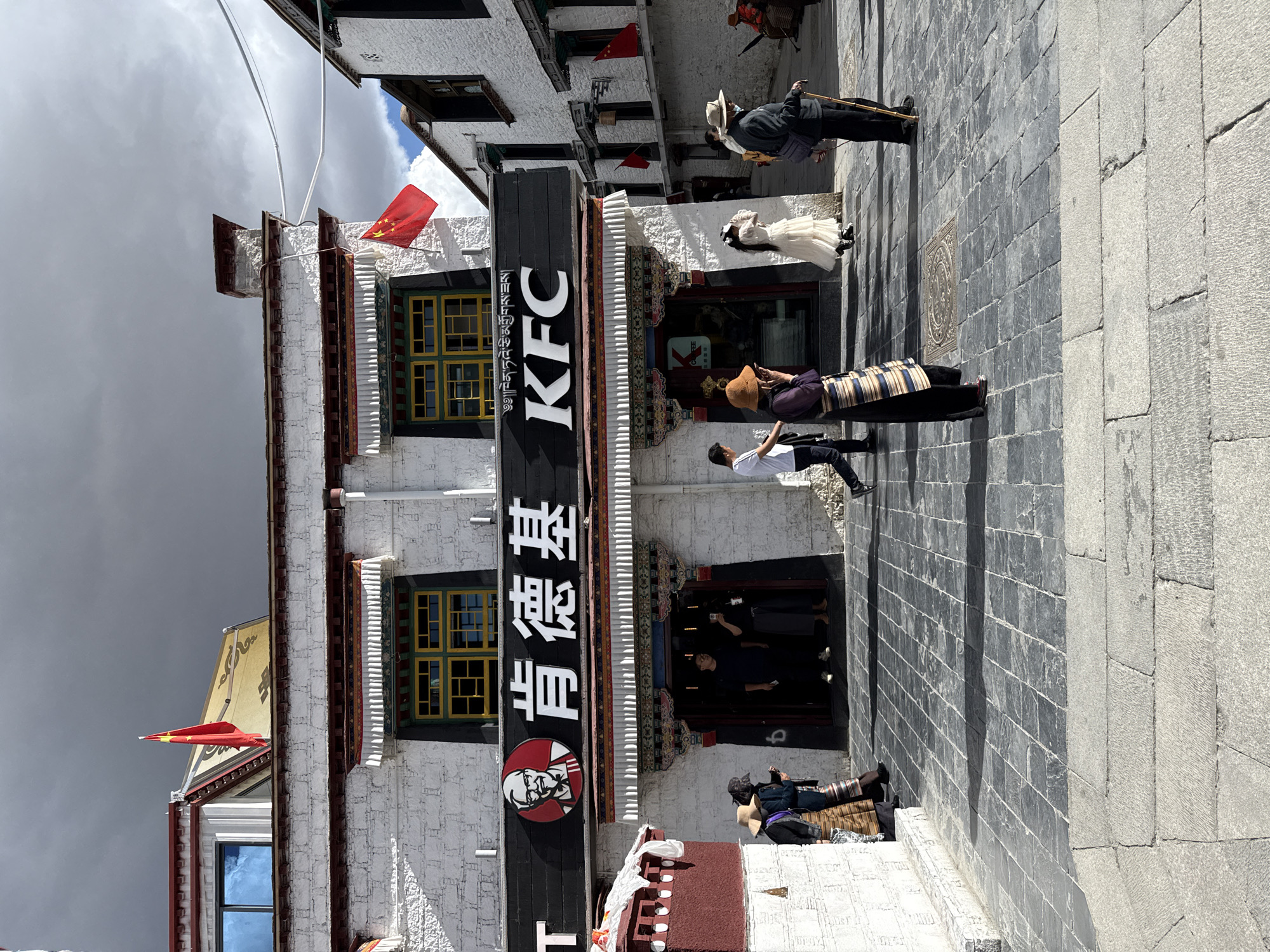 This vertical image captures a lively street scene in Chengguan District, China, featuring a prominent KFC restaurant housed within a building that blends modern commerce with traditional architecture. The sky above is largely overcast, though patches of blue are visible, indicating a daytime setting.
The main subject is a multi-story building with distinct white textured walls, characteristic of traditional Tibetan-style architecture, featuring intricate dark wooden window frames with yellow and green panels, and decorative eaves. A large black sign runs vertically down the building's facade, boldly displaying "KFC" in white English letters and "肯德基" (Kěndéjī), the Chinese name for KFC, along with the iconic Colonel Sanders logo. Several Chinese national flags (red with five yellow stars) are affixed to the building's upper sections and rooflines.
On the stone-paved street below, several people are visible. Near the KFC entrance, a person in a white dress and a light-colored hat stands, possibly a tourist or a local in traditional attire. Another individual in what appears to be traditional Tibetan clothing, featuring a striped apron or skirt and a dark hat, stands closer to the foreground. A person in a white t-shirt and dark pants is seen holding a device, possibly taking a photograph or viewing content. Further down the street, an individual in a dark jacket and hat walks with a cane. Inside the well-lit entrance of the KFC, figures can be seen, suggesting customers. To the lower left, a person is seated on the ground with a bag. The street and adjacent buildings are paved with large, irregularly cut stone blocks, and one side building features a decorative circular emblem on its stone wall.