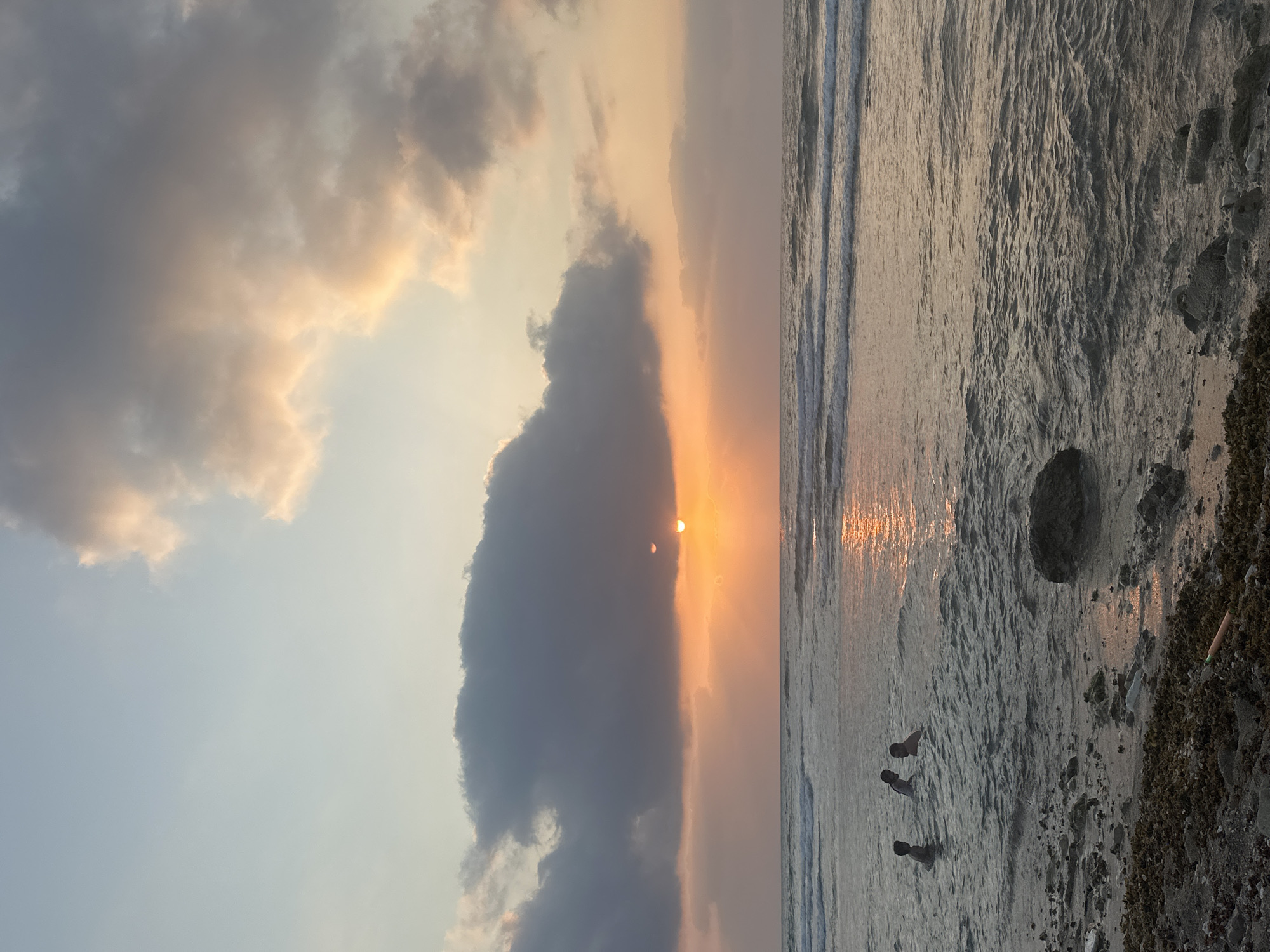 The image depicts a sunset over the Mediterranean Sea in Haifa, Israel. Three individuals are visible wading in the shallow water near the shore. The sky is a mixture of soft blues and oranges, with clouds tinged with sunset colors. The sun is partially obscured by a large, dark cloud, casting a shimmering reflection on the water. The shoreline is rocky and covered with debris, including seaweed and scattered rocks. The scene appears to be a tranquil evening at the beach.