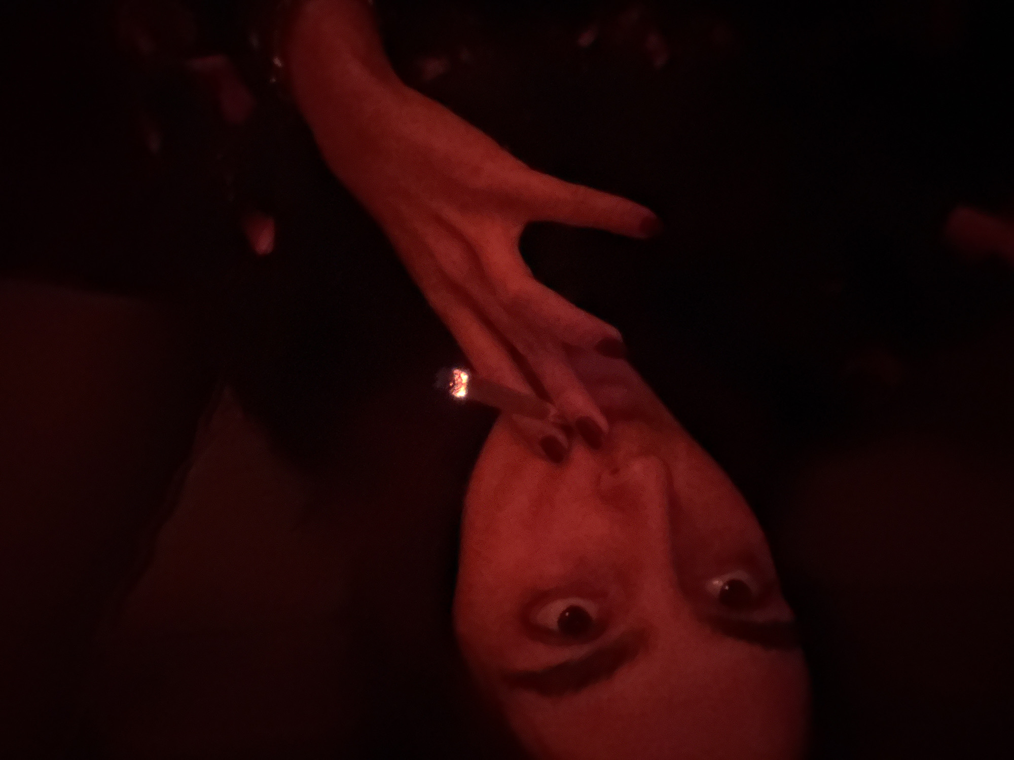 A close-up, rotated portrait captures a person in a dimly lit, predominantly red environment. The subject's face, seen from the eyes down to the chin, and one hand are visible. The person looks directly at the camera with wide, intense eyes. A lit cigarette, glowing bright white and orange at its tip, is held between the fingers of their right hand, positioned near their mouth. The fingers show dark nail polish.
The scene is dominated by a strong red artificial light source, casting a deep crimson glow over the subject's skin and hand, while the background remains almost entirely black, save for some faint, indistinct shapes in the upper left. The overall atmosphere is dark, dramatic, and perhaps mysterious, suggesting a nocturnal setting, possibly within a club or bar in Mexico City, Mexico. No visible text is present.