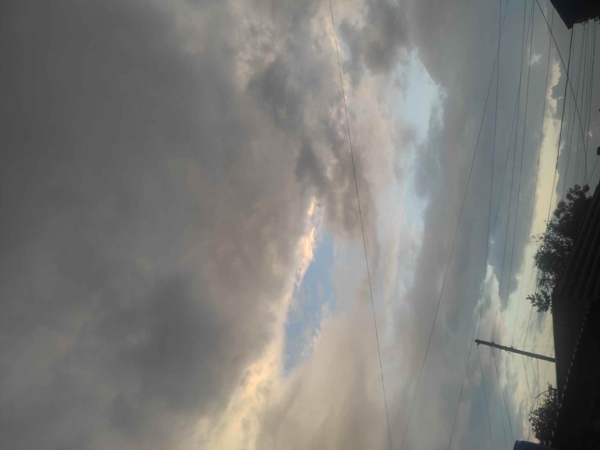 The image depicts an overcast sky with patches of blue visible through breaks in the clouds. The lower portion of the image shows the silhouette of rooftops, trees, and utility poles with power lines. The scene appears to be a residential area in Shakhtinsk, Kazakhstan, given the architecture and the presence of power lines typical of such locations. The lighting suggests it might be late afternoon or early evening. There are no people or discernible actions in the image, focusing solely on the environment and atmospheric conditions.