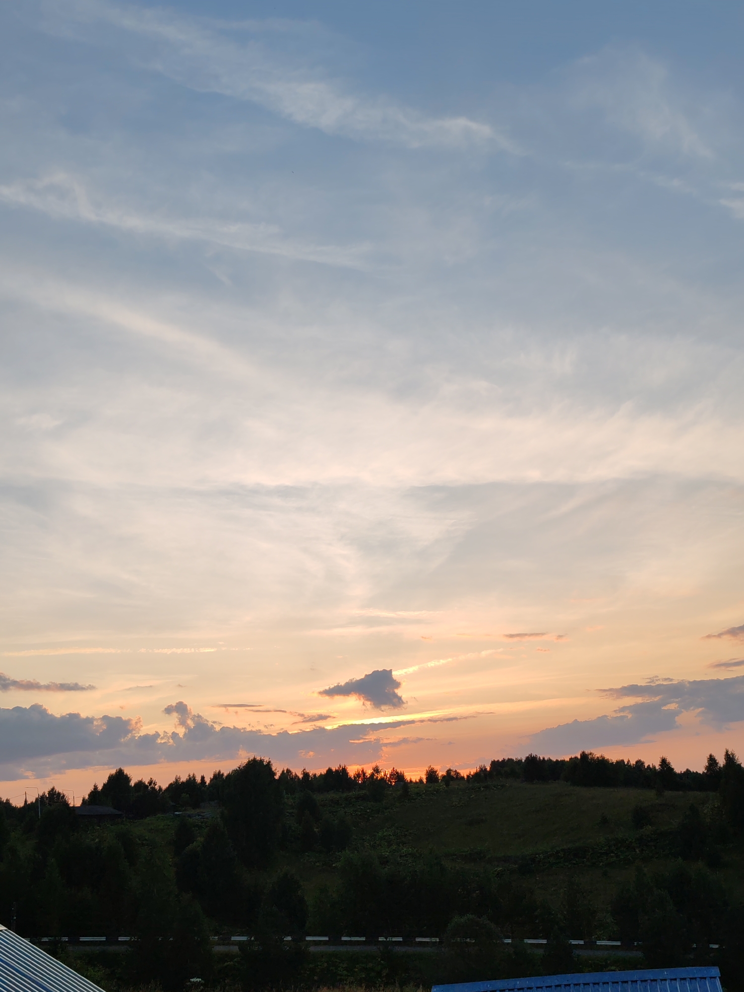 The image depicts a rural landscape at sunset, likely in or near Чухлэм, Russia. The sky is a gradient of blue and orange, with wispy clouds scattered across it. The sun is low on the horizon, casting a warm glow over the scene. In the foreground, a rolling green hill is covered with dense trees. A small building, possibly a house, is visible nestled among the trees. A road runs along the base of the hill, marked by a white barrier. In the bottom left corner, a section of a corrugated metal roof is visible, suggesting a man-made structure or enclosure. The overall impression is one of tranquility and natural beauty. There are no people or animals visible in the image.