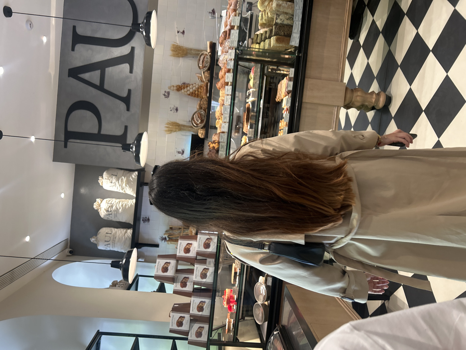 The image captures an indoor scene at a "PAUL" bakery, located in Yerevan, Armenia. The primary subject is a person, seen from behind, with long, dark brown hair. They are wearing a light beige or tan trench coat and carrying a small black shoulder bag, with one hand holding a mobile phone.
The setting is a well-lit bakery interior with a modern, clean design. A large, dark grey sign with the prominent black letters "PAUL" is visible on the upper wall. Below this, there are shelves displaying sacks, some also bearing the "PAUL" brand, likely containing flour or other ingredients. On the right, a display case showcases a variety of baked goods, including croissants and other pastries, alongside what appear to be cheeses. Further down, another display cabinet holds boxes labeled "PANETTONE." The walls are white or light-colored, with a section of white subway tiles visible behind the pastry display. The floor features a distinct black and white checkerboard pattern.
The person appears to be walking through or browsing the bakery, possibly heading towards a counter or another display. The overall ambiance suggests a busy, inviting bakery environment, typical of the PAUL chain. The lighting is bright, provided by overhead spotlights and black pendant lamps. No specific time of day or outdoor weather is discernible.
