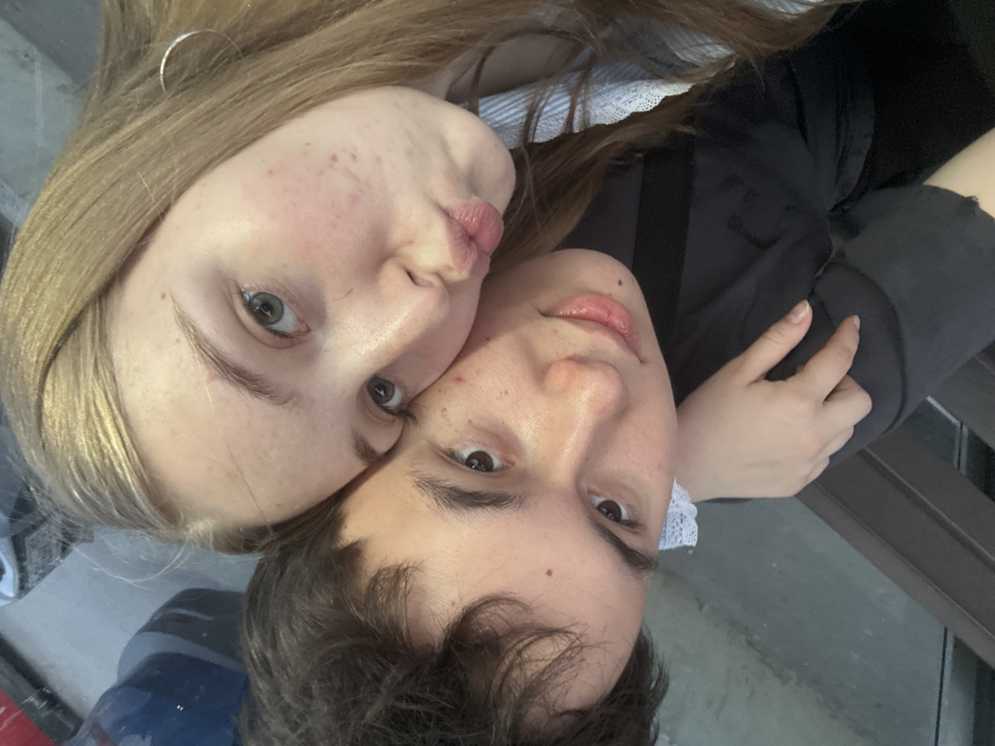 The image shows two people in an embrace, likely a couple. The person on the left has light hair and pale skin, with noticeable blemishes on their face. They are wearing a gold hoop earring and a top with a lacy collar is visible. Their lips are pursed, as if posing for a kiss. The person on the right has dark hair and appears to be leaning their head against the other person. Both are looking directly at the camera. The location appears to be outdoors, with a gray surface beneath them. The angle of the photo suggests it's a selfie. The city of Perm, Russia is not discernable from the image.