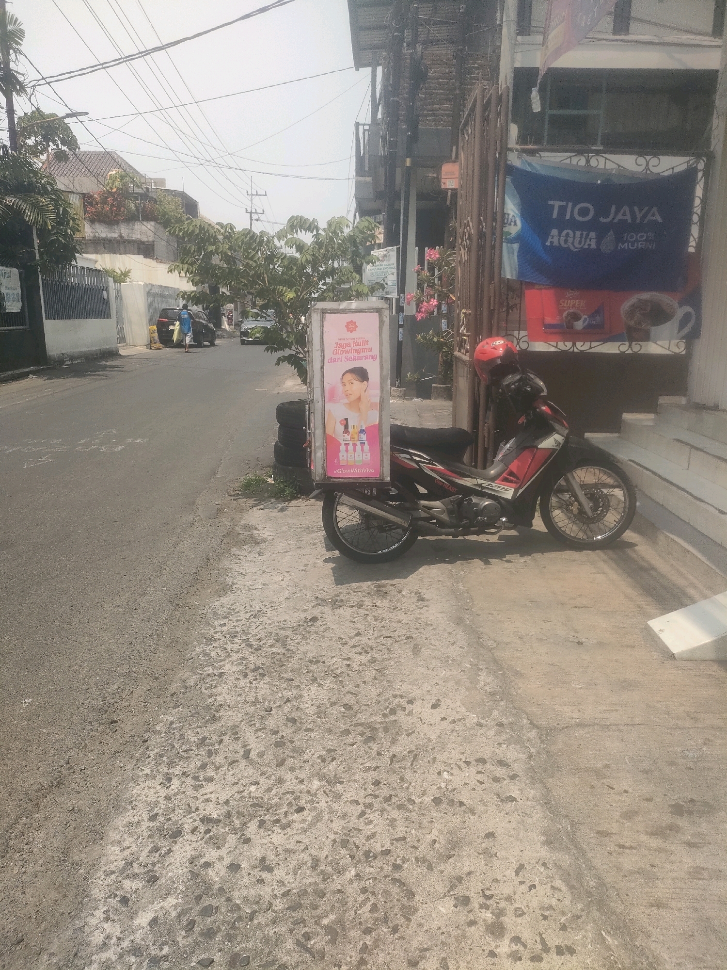 A side view of a motorcycle with a promotional sign is parked on a sidewalk next to a street. The motorcycle is red and black and has a red helmet on the handlebars. The promotional sign is advertising beauty products. In the background, there is a building with a blue banner that reads "TIO JAYA AQUA 100% MURNI". A person in a blue shirt is standing on the street next to a black car. The street is paved and has some cracks and debris. The sidewalk is concrete and also has some debris. The sky is hazy and overcast. This scene is in Surabaya, Indonesia.