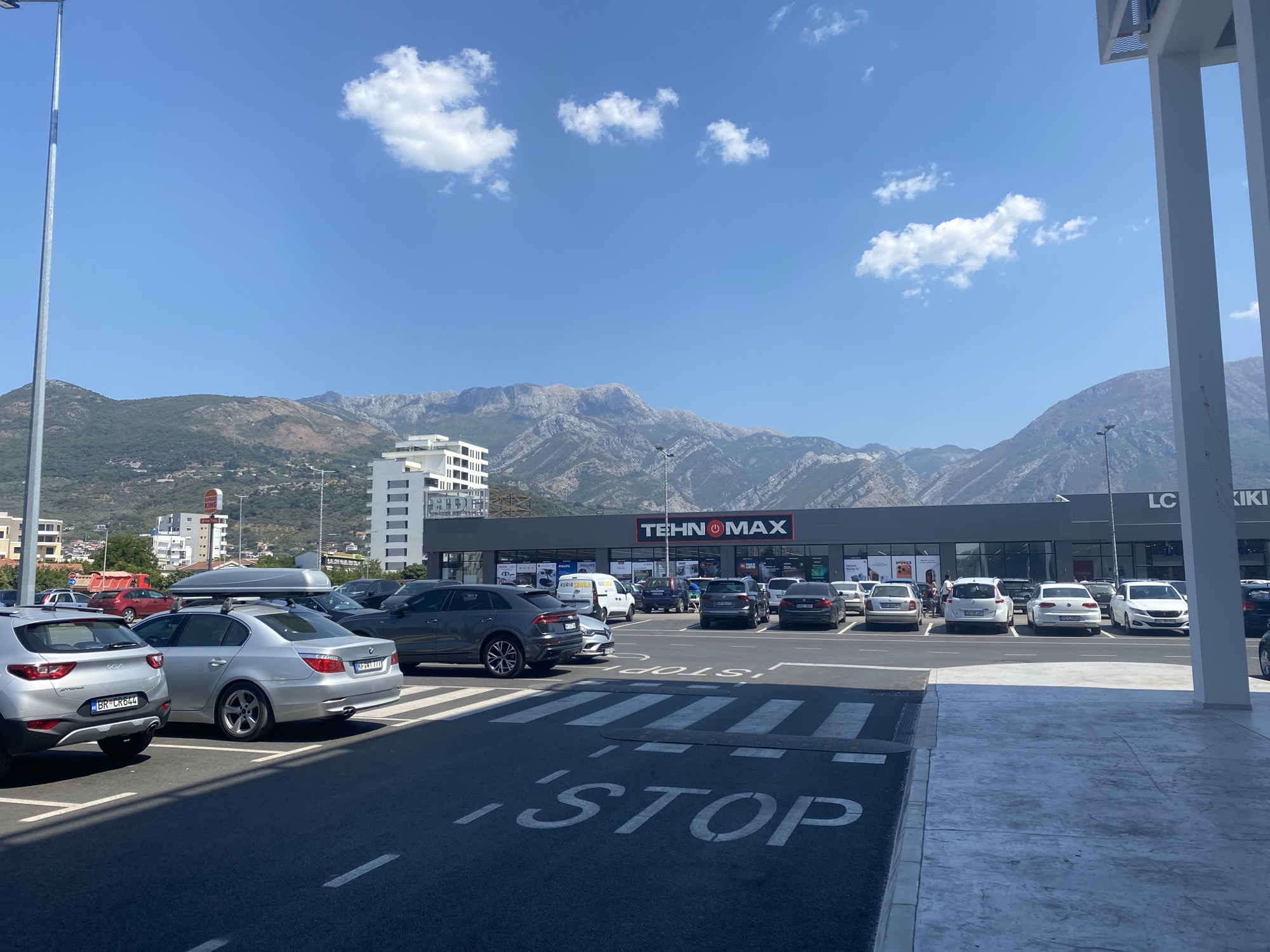 The image shows a parking lot in front of a commercial building with the "TEHNOMAX" logo prominently displayed. The building appears to be a retail store, possibly electronics or home goods, with several cars parked in front of it. The scene is outdoors, with a clear blue sky and mountains in the background. There are several cars parked in the lot, some with their license plates visible. One car has a roof box, suggesting travel. A street sign with "STOP" is painted on the asphalt in the foreground. The overall environment suggests a sunny day in a city or town. The location is identified as Bjeliši, Montenegro.