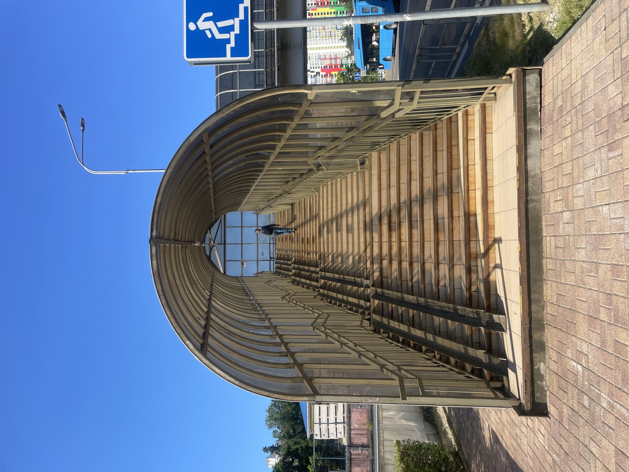 The image displays a long, covered pedestrian crossing or underpass entrance in Sochi, Russia, captured on a clear, sunny day. The structure features a large, arched, translucent roof supported by a light-colored metal framework, with steps leading upwards. A single person, dressed in dark clothing, is visible walking up these steps inside the covered walkway, illuminated by bright sunlight which casts strong, sharp shadows from the framework onto the steps.
The ground leading up to the structure is paved with reddish-brown bricks or tiles. To the left, a tall street lamp with two fixtures extends into a vibrant, cloudless blue sky. Above the entrance, a blue sign with white pictograms indicates a pedestrian facility, showing a figure ascending/descending stairs alongside a wheelchair symbol, denoting accessibility.
In the background, visible through and beyond the pedestrian crossing, is a multi-lane road where a blue bus is driving. Further back, urban buildings are discernible, including one with a striking facade featuring vertical stripes of various bright colors (red, orange, yellow, green, blue). Scattered trees and bushes add touches of greenery to the urban landscape. The overall scene suggests a well-maintained public space under bright, daytime conditions.