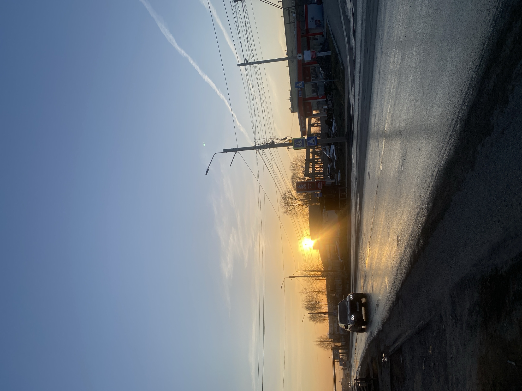 The image shows a sunrise in Chaikovsky, Russia, along a wet street. The sky is blue with contrails above, transitioning to an orange glow near the horizon. A black car with its headlights on drives toward the viewer. Buildings, streetlights, and power lines line the street, and a gas station is visible in the distance. The road surface is reflective due to moisture.