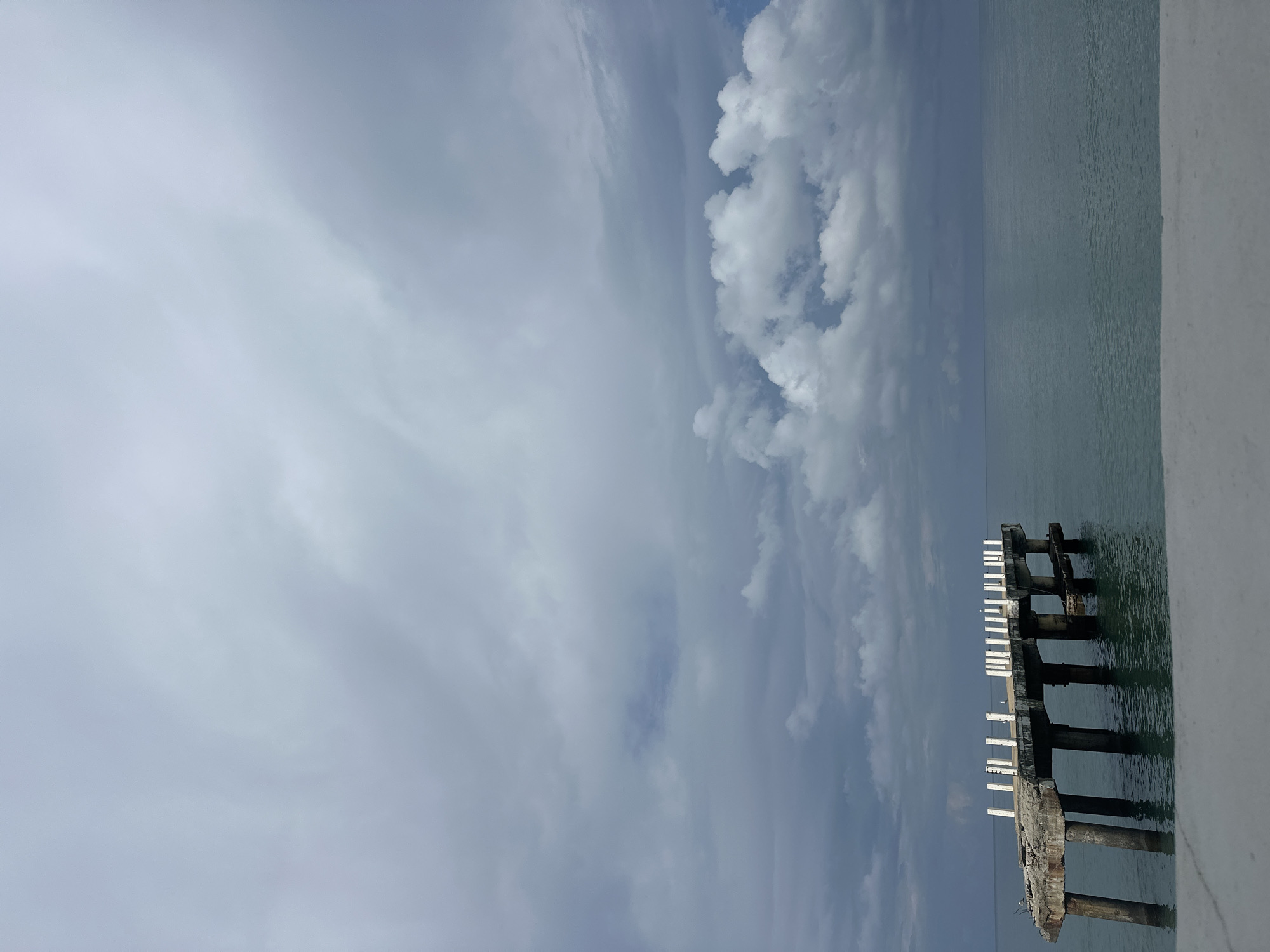 The image depicts a partially ruined pier extending into a calm, teal-colored sea under a cloudy sky. The pier, constructed of concrete and supported by thick pillars, shows signs of significant damage and decay, with sections of the platform missing and broken railings. White vertical posts line the top of the remaining sections of the pier. The sky is filled with a mix of dark gray and lighter white clouds, suggesting overcast or impending weather. The sea is still, with subtle ripples visible on its surface. In the foreground, a light-colored, solid structure, possibly a wall or railing, occupies the bottom portion of the frame. The scene is devoid of any people or animals. The overall atmosphere is one of abandonment and natural decay. This pier is located on the coast of Batumi, Georgia, a popular Black Sea resort city.