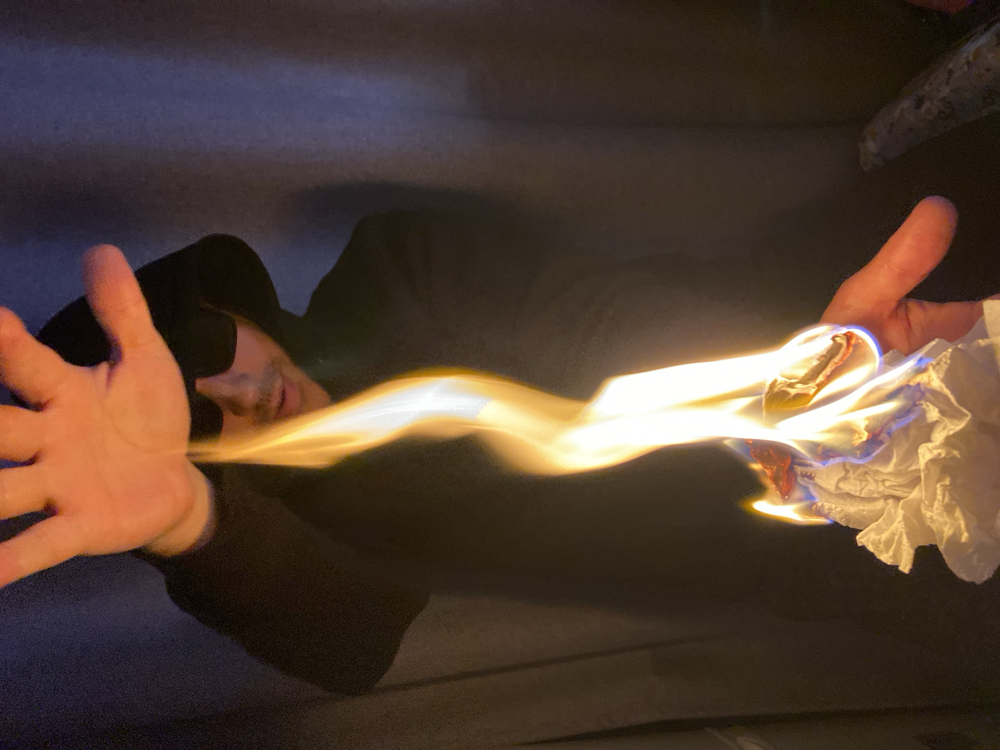 A person wearing a dark hooded sweatshirt and sunglasses is lying down. Between their outstretched hands is a stream of fire, emanating from a wad of crumpled paper/tissue held in the person's right hand. The background appears to be an interior setting. Location: Unknown, Unknown.