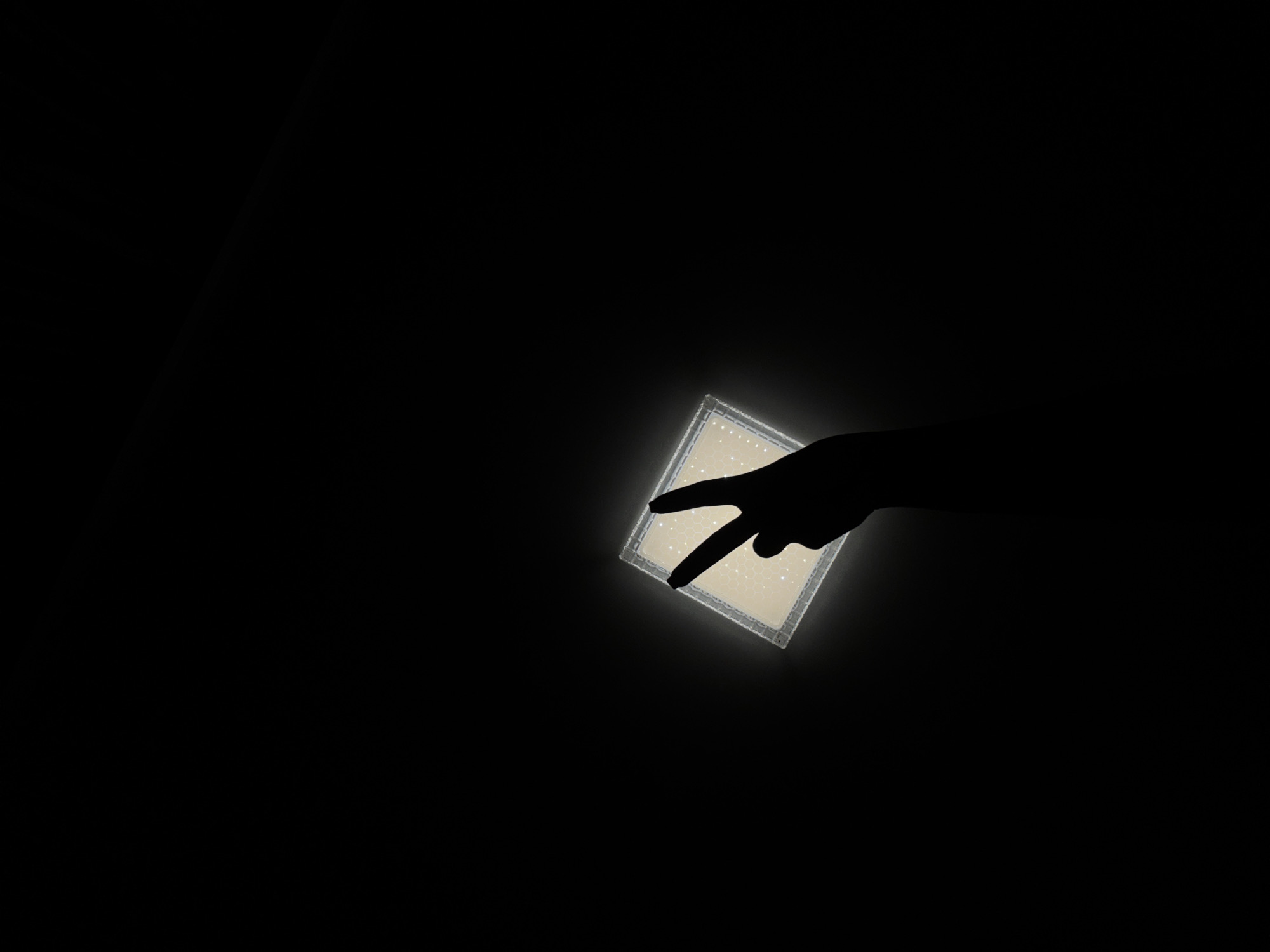 A hand silhouette holds a glowing square object, casting a peace sign shadow. The background is completely black. The location Марковский, Russia, is irrelevant.